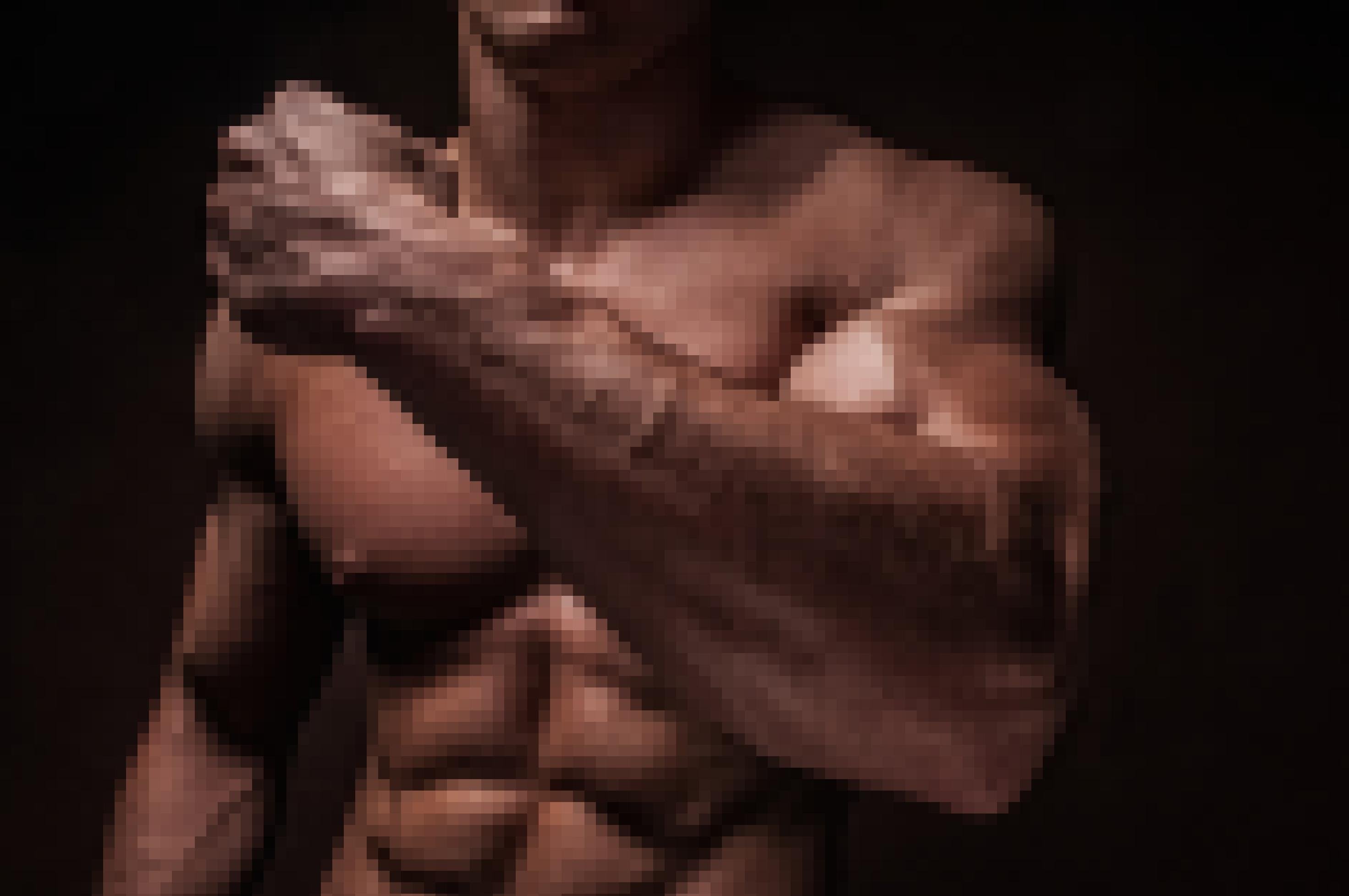 Zu sehen ist vor dunklem Hintergrund der angeleuchtete, braune, nackte Oberkörper eines Mannes mit Waschbrettbauch und ausgeprägter Brustmuskulatur. Er hält seinen linken Arm angewinkelt vor sich und zeigt dabei seinen riesigen Bizeps.