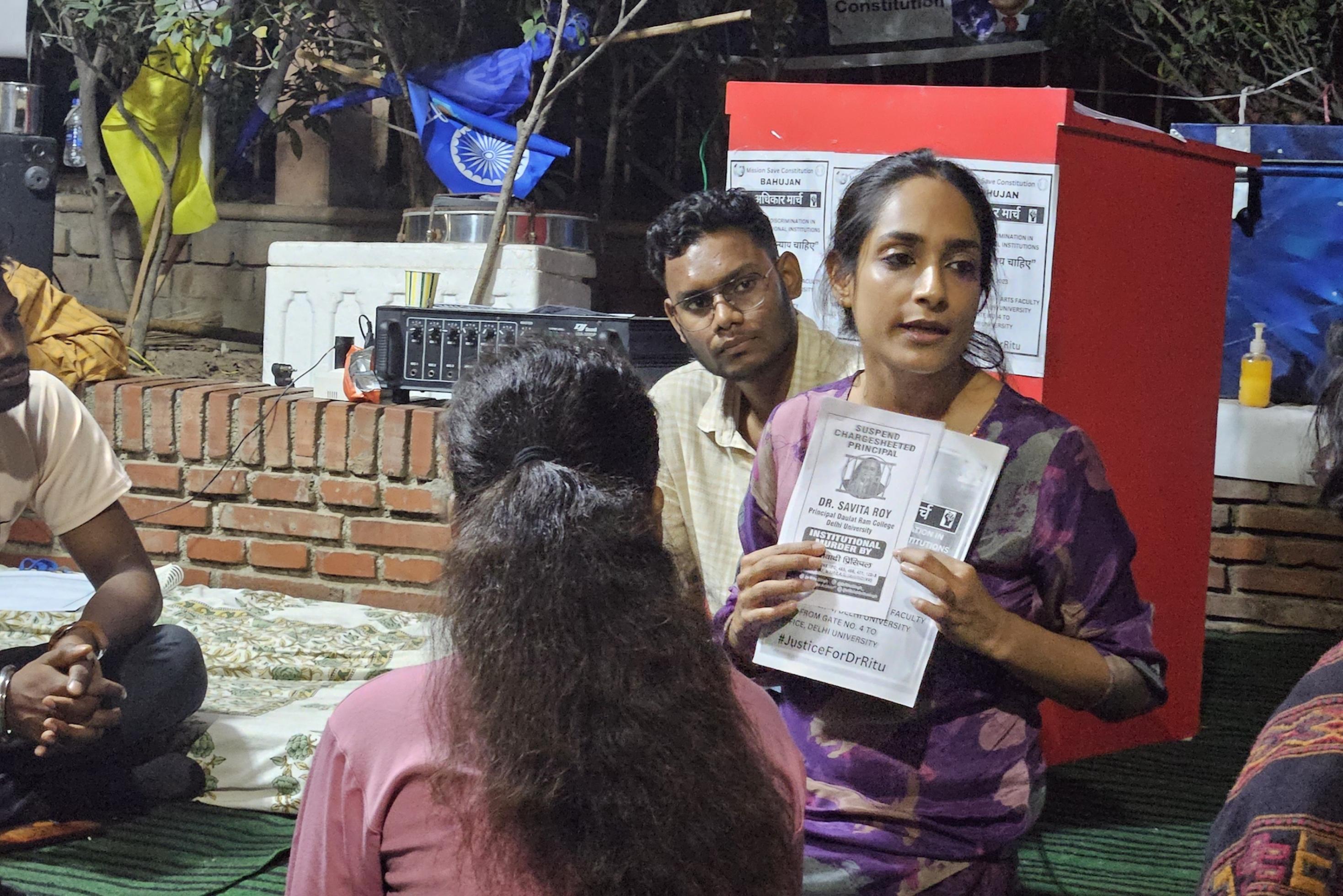 Eine indische aussehende Frau sitzt umringt von drei Studierenden auf dem Boden und zeigt auf ein Papier in ihrer Hand.
