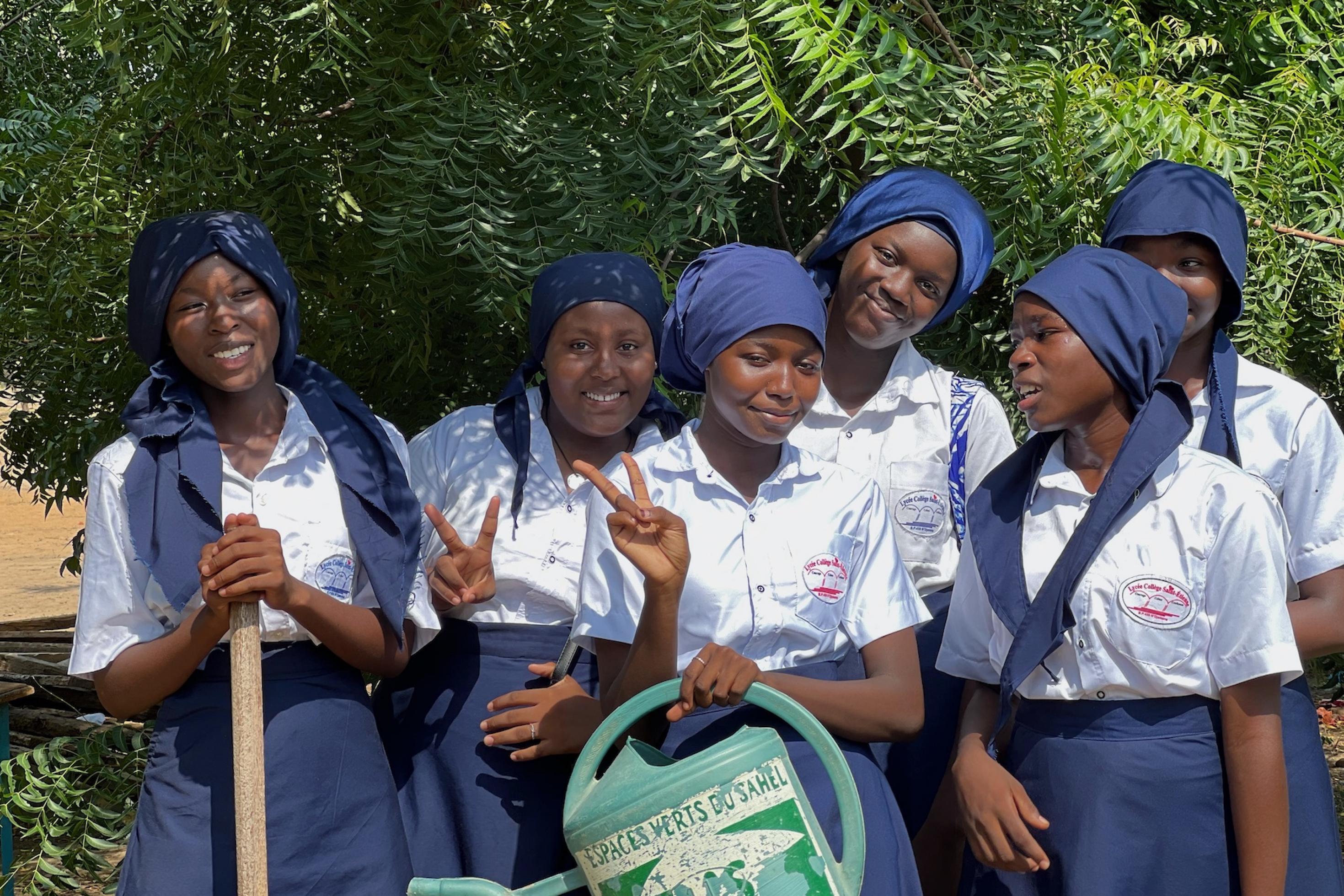 Sechs junge Frauen aus dem Tschad stehen in blauweißer Schuluniform vor einer grünen Hecke und blicken lachend in die Kamera. Eine hält eine Gießkanne in der Hand.