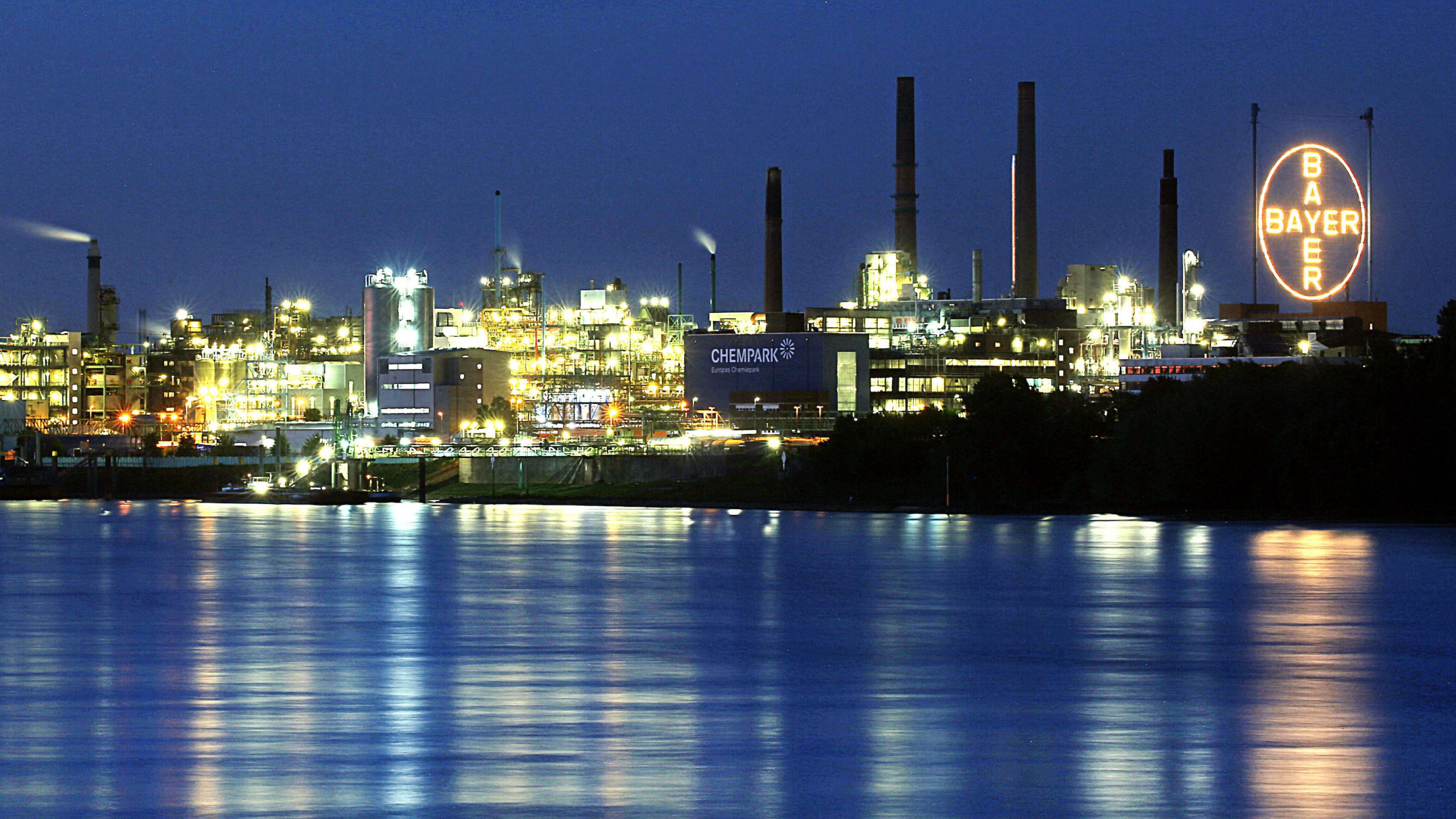 Nächtliche Aufnahme vom Rhein aus gesehen, der Fluss im Vordergrund dahinter die hell beleuchtete Chemieanlage mit dem großen Bayer-Logo.