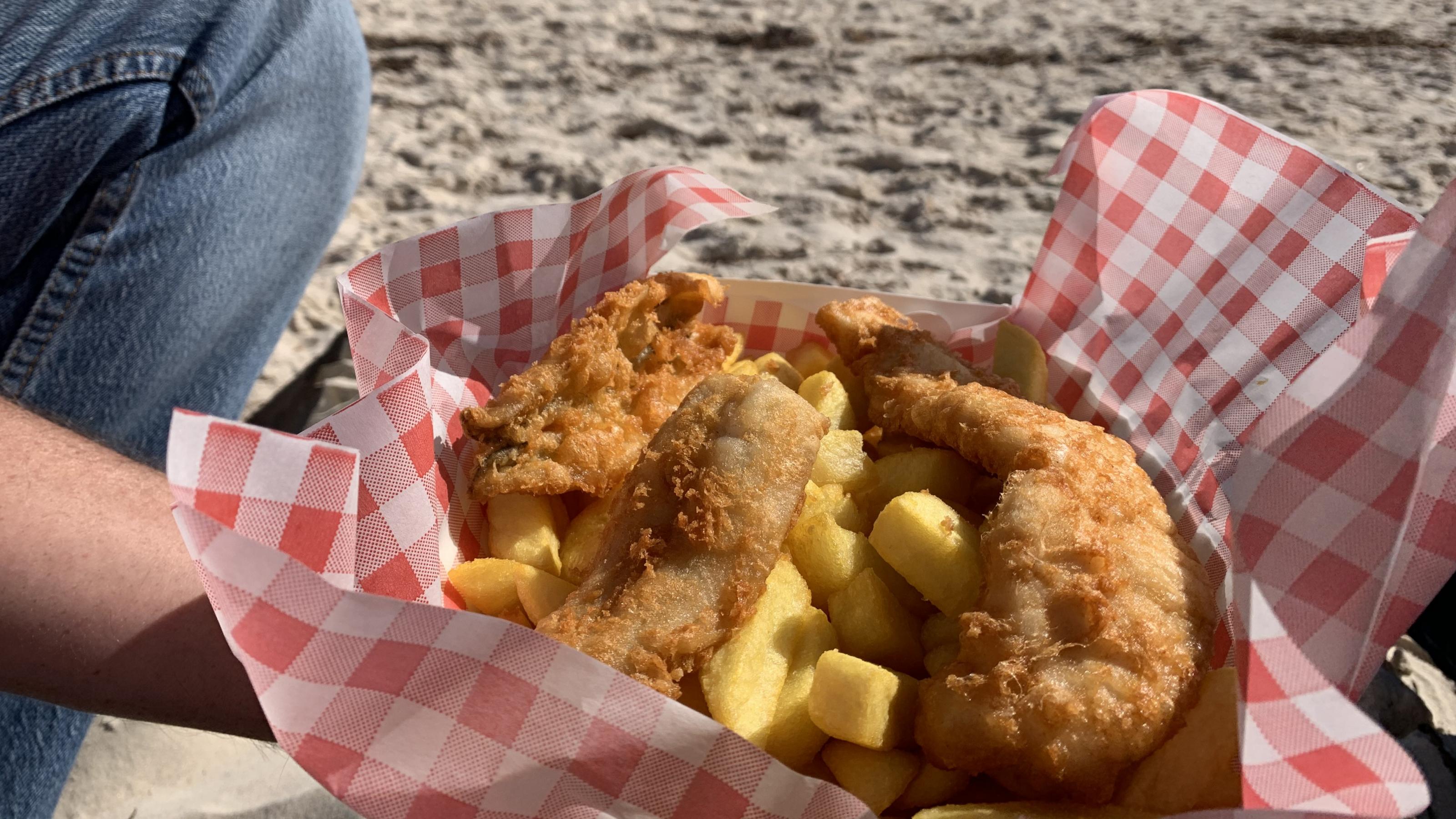 Das Foto zeigt eine Portion Fisch & Chips, die an einem Strand verspeist wird.