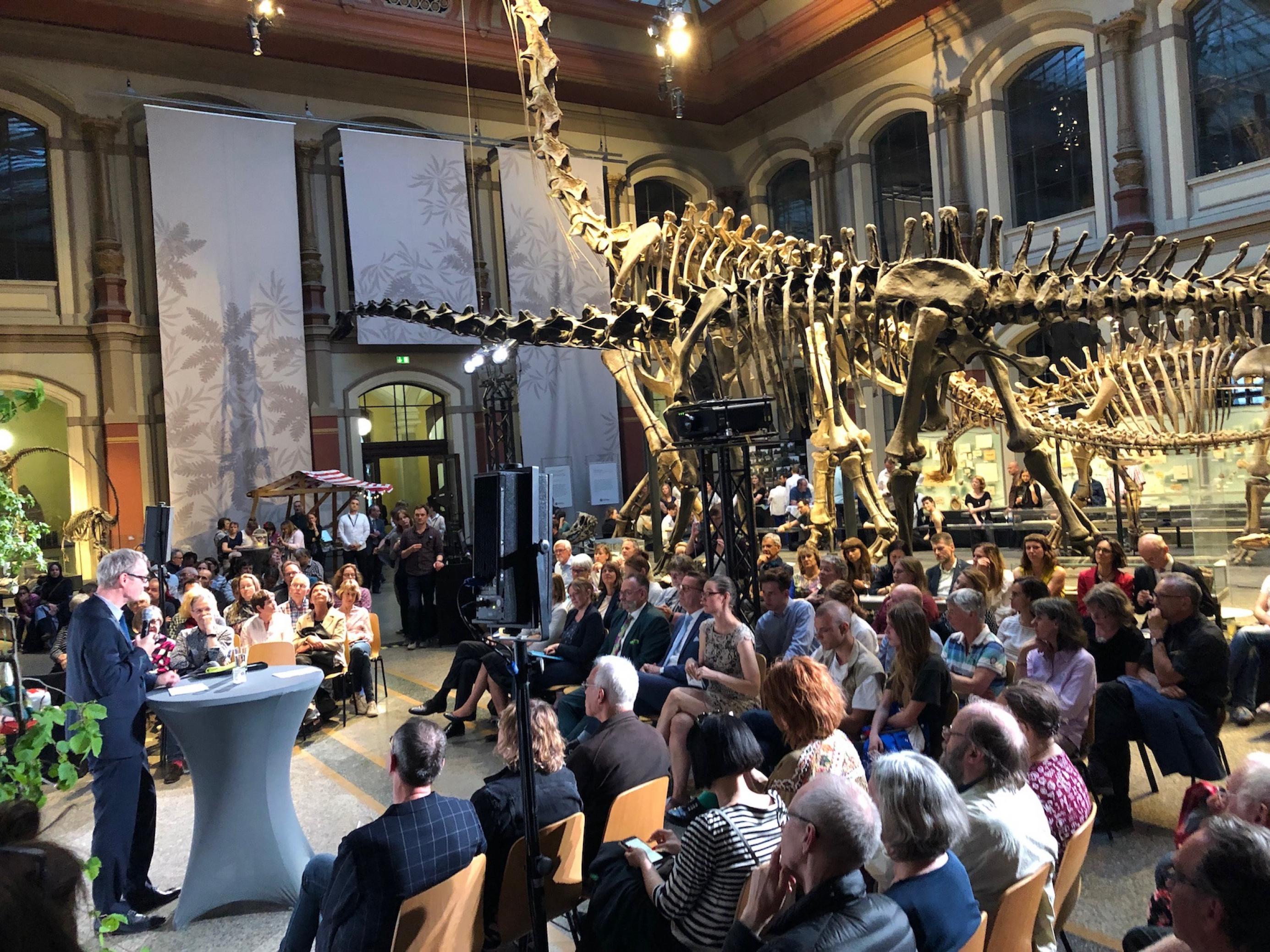 Viele Menschen sitzen um ein Redepult herum. Daran steht ein Mann im Anzug. Zwischen den Zuschauern befindet sich ein sehr großes Skelett eines Dinosauriers.