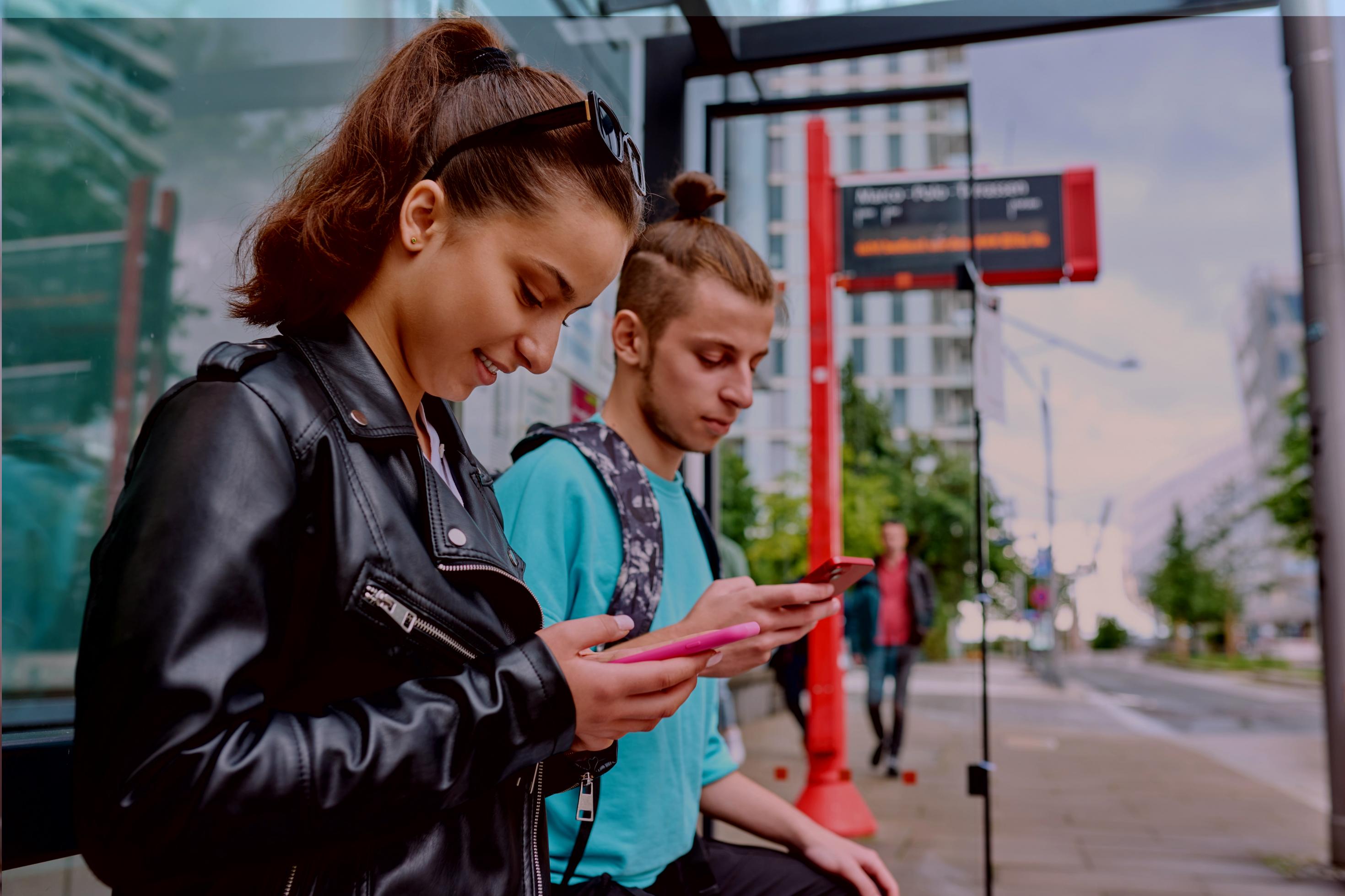 Zwei gut gelaunte junge Menschen schauen an einer Haltestelle auf ihre Handys.