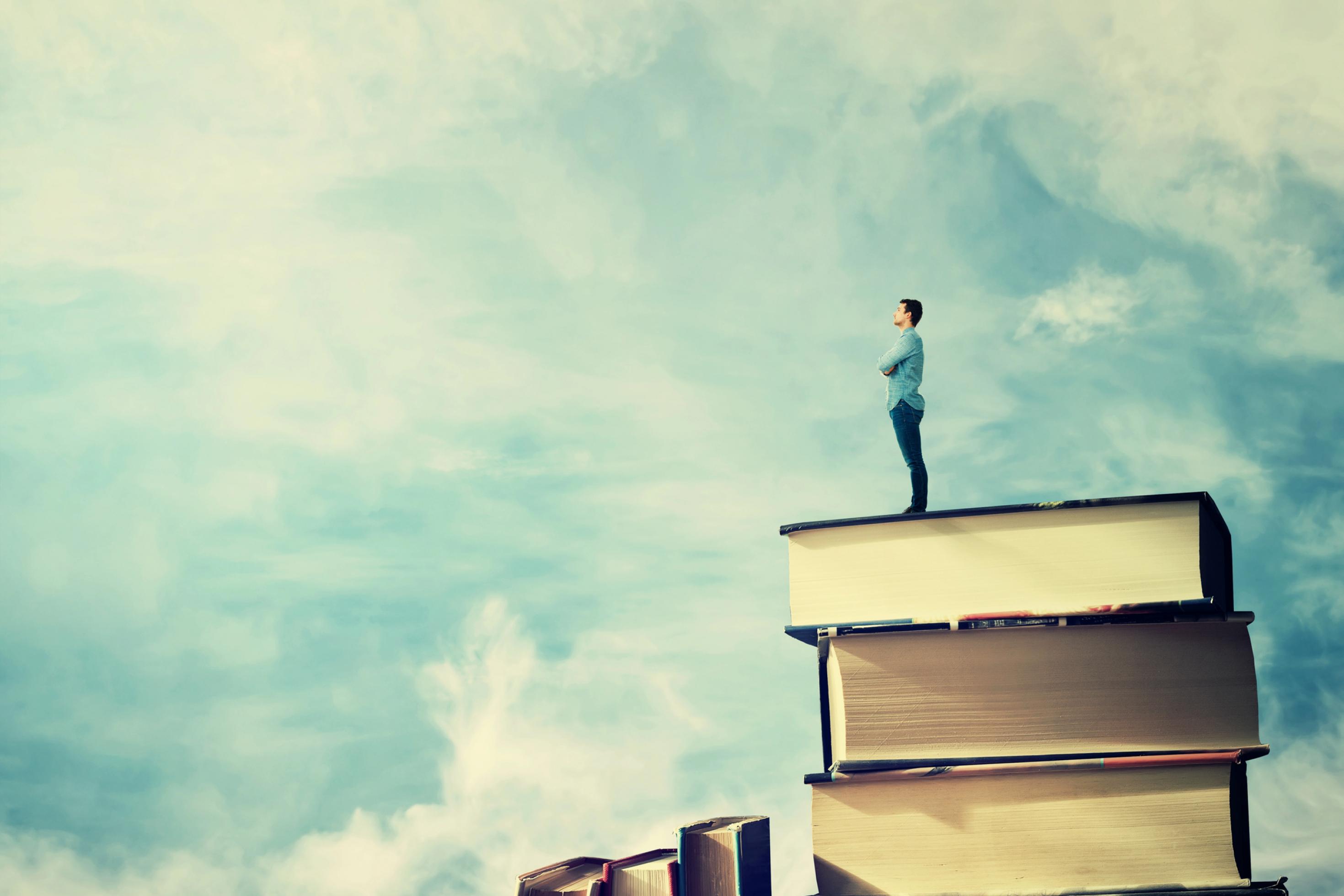 Fotomontage, in der ein Mann in legerer Kleidung auf einem Stapel Bücher steht und in einen blauen Himmel mit einigen weißen Wölkchen blickt.