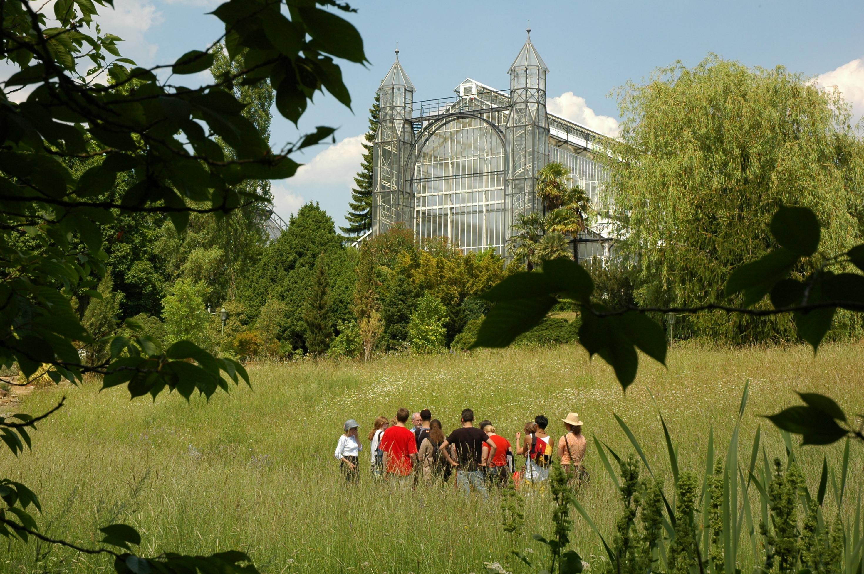 Das Bild zeigt ein Treibhaus des Botanischen Gartens Berlin, das sogenannte Mittelmeerhaus, eingebettet in  die parkartige Landschaft. Dort steht eine Gruppe junger Menschen mit einem Dozenten.