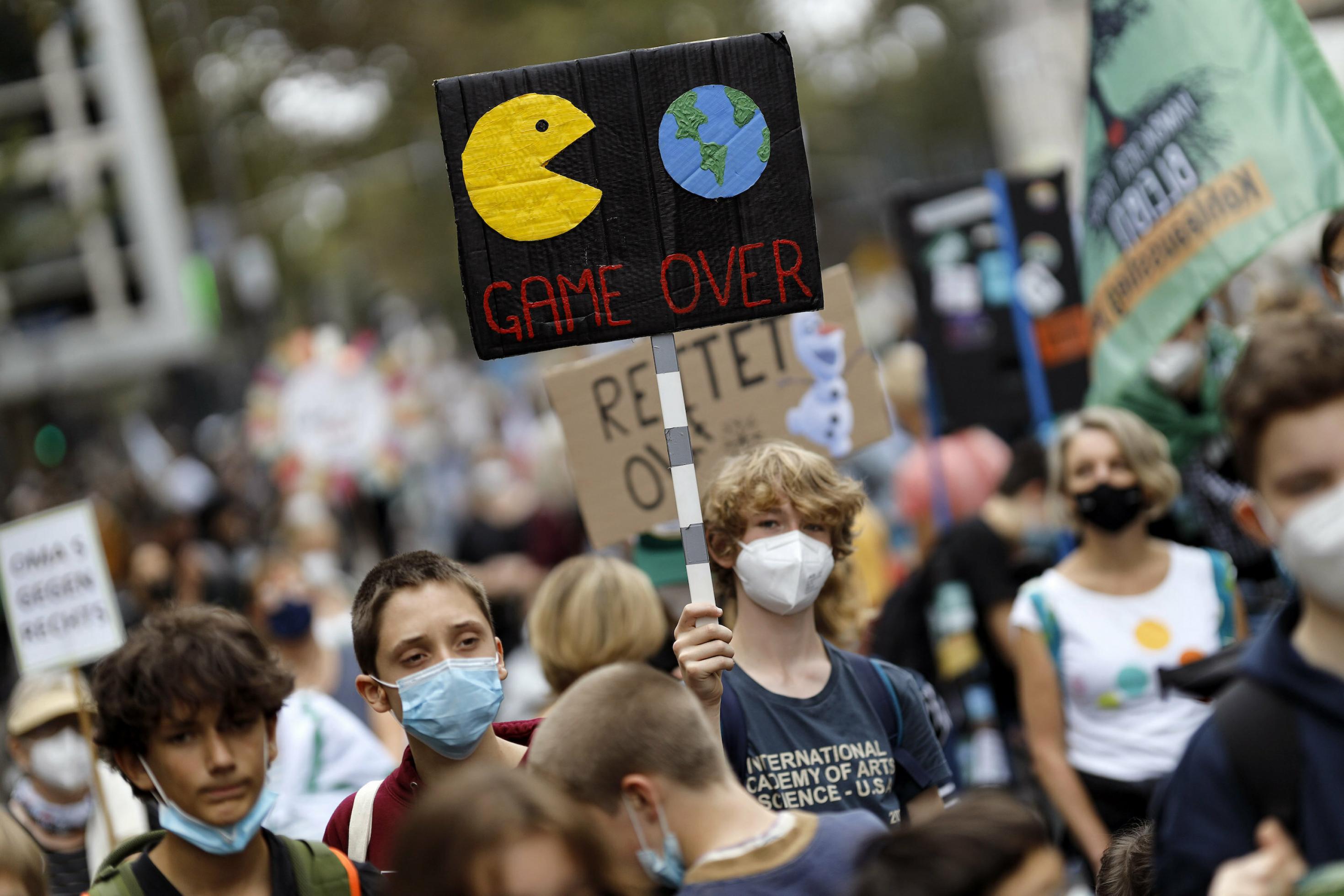 Ein junger Demonstrant hält ein Schild hoch, auf dem ein Packman-Monster droht, die Erde aufzufressen. Beschriftung: „Game over“.