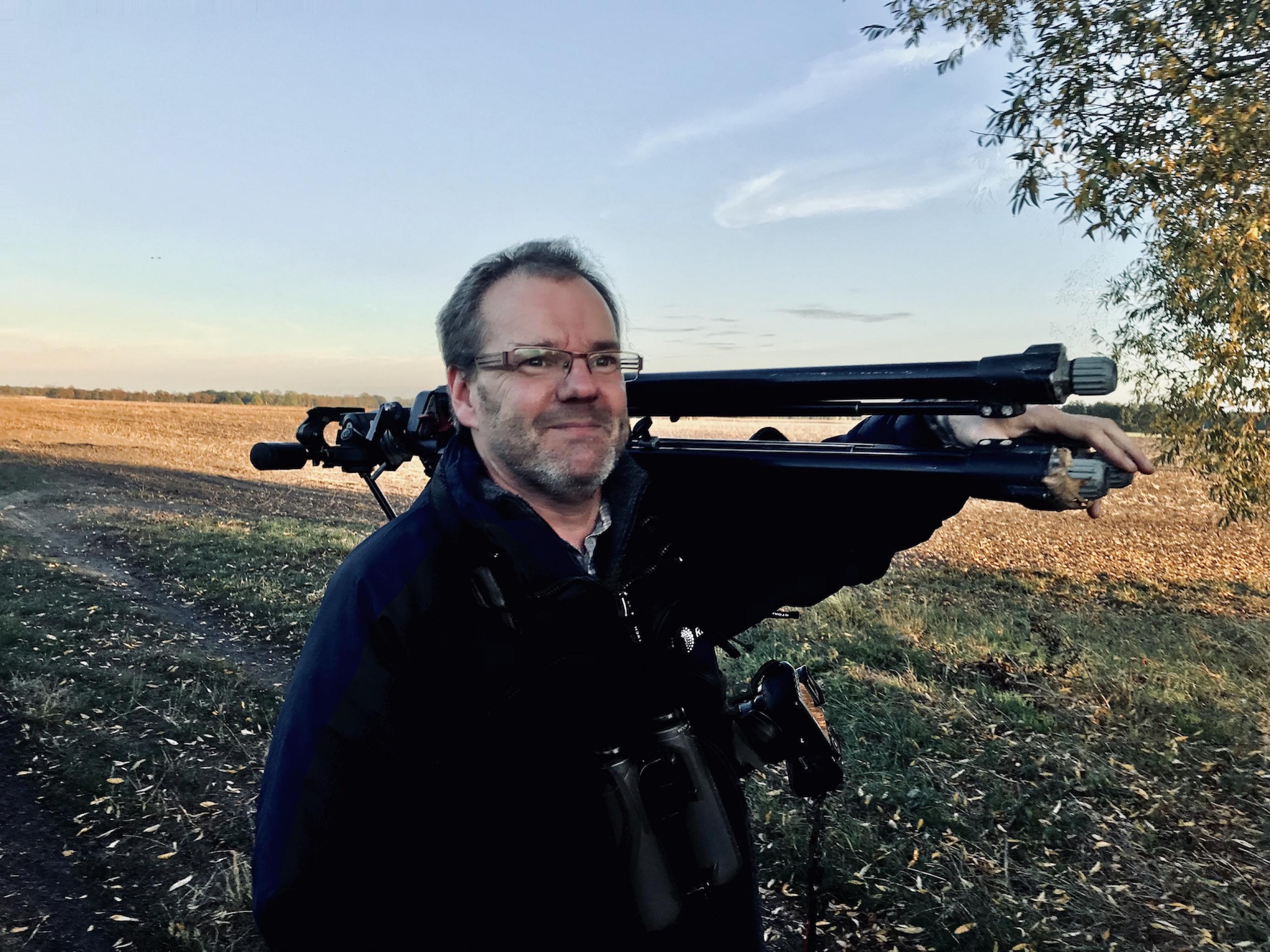 Portrait von Thomas Krumenacker auf einem Feld. Er schultert ein Stativ und trägt ein Fernglas um den Hals.