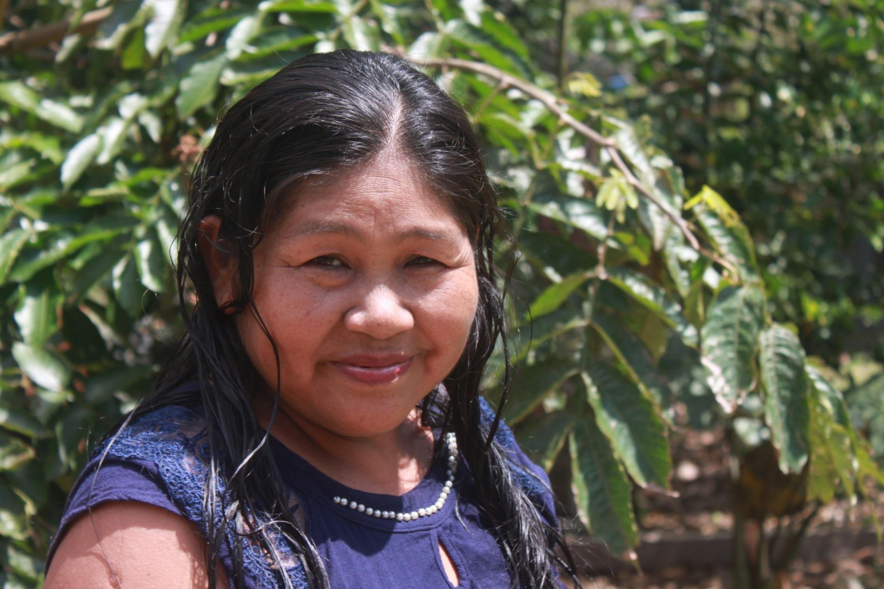Brustbild einer rund 50-jährigen indigenen Frau mit langen schwarzen Haaren. Im Hintergrund grünes Buschwerk.