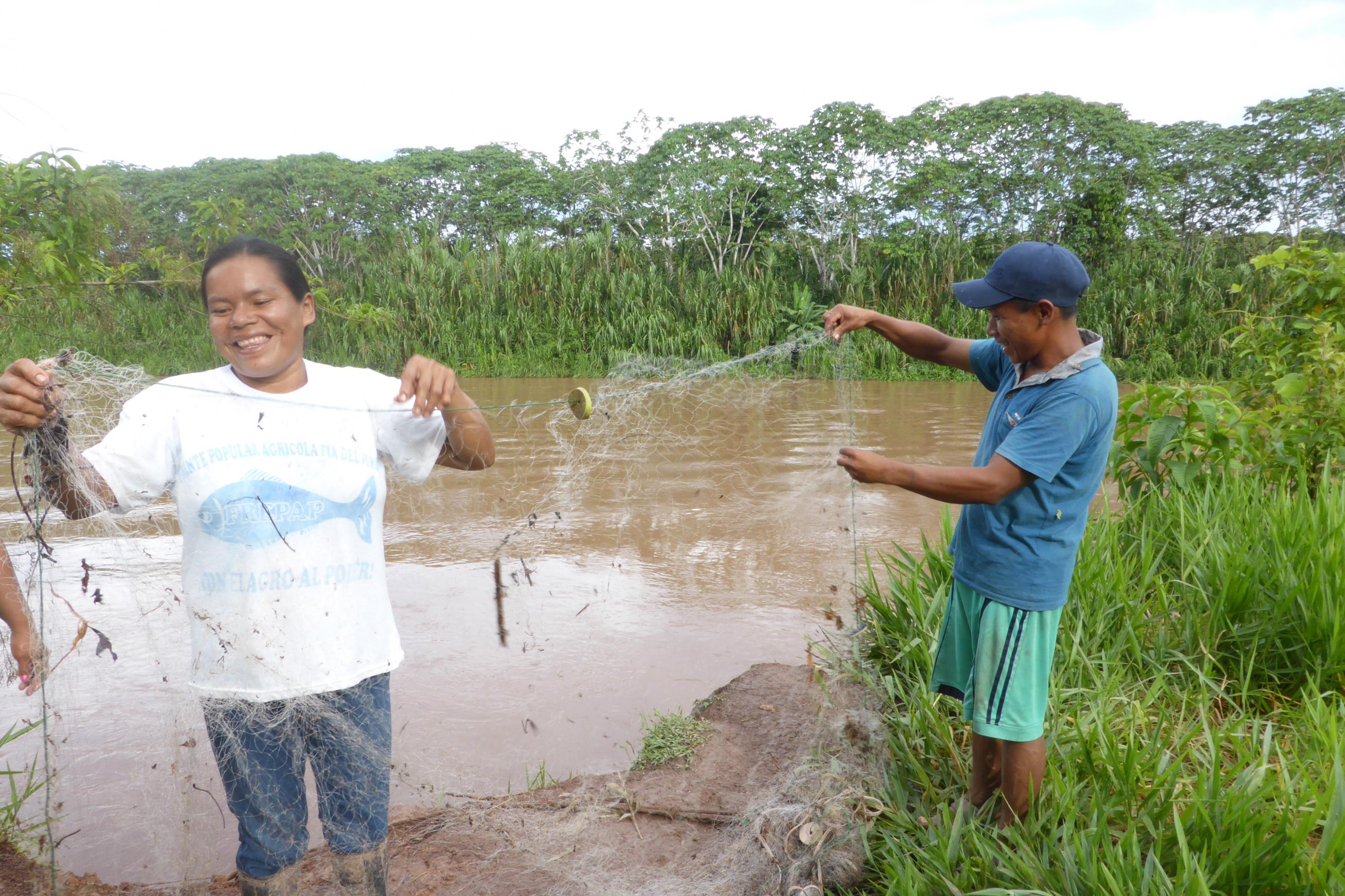 Eine Frau vom Volk der Ashaninka und ein Mann stehen am Ufer eines Flusses und säubern ein Fischernetz von Steinchen, Blättern und Ästen, die beim Fischen dort hängen geblieben sind.