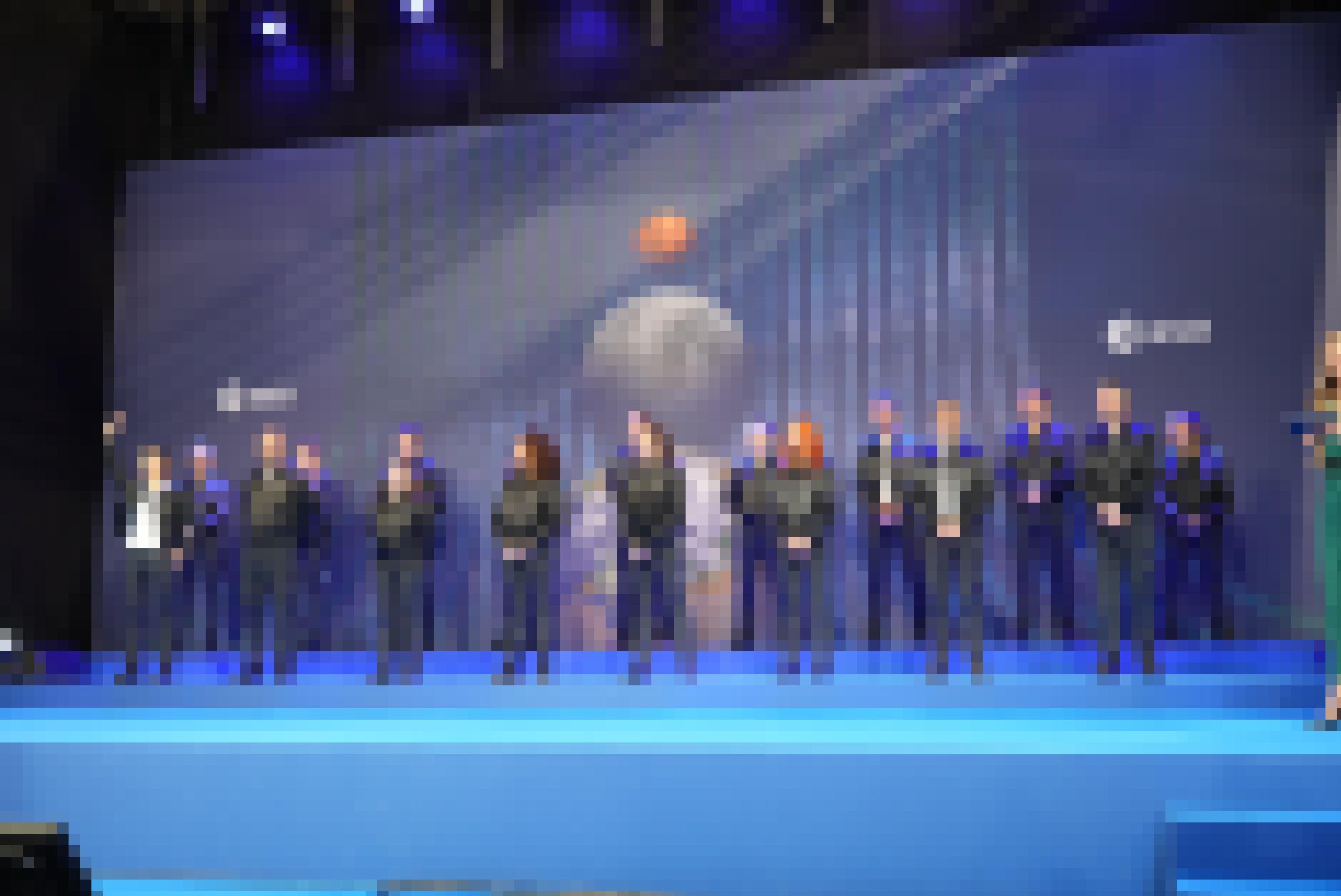 Auf einer blauen Bühne stehen siebzehn Personen. Im Hintergrund ist ein Bild mit Perspektive auf Erde, Mond und Mars eingeblendet.