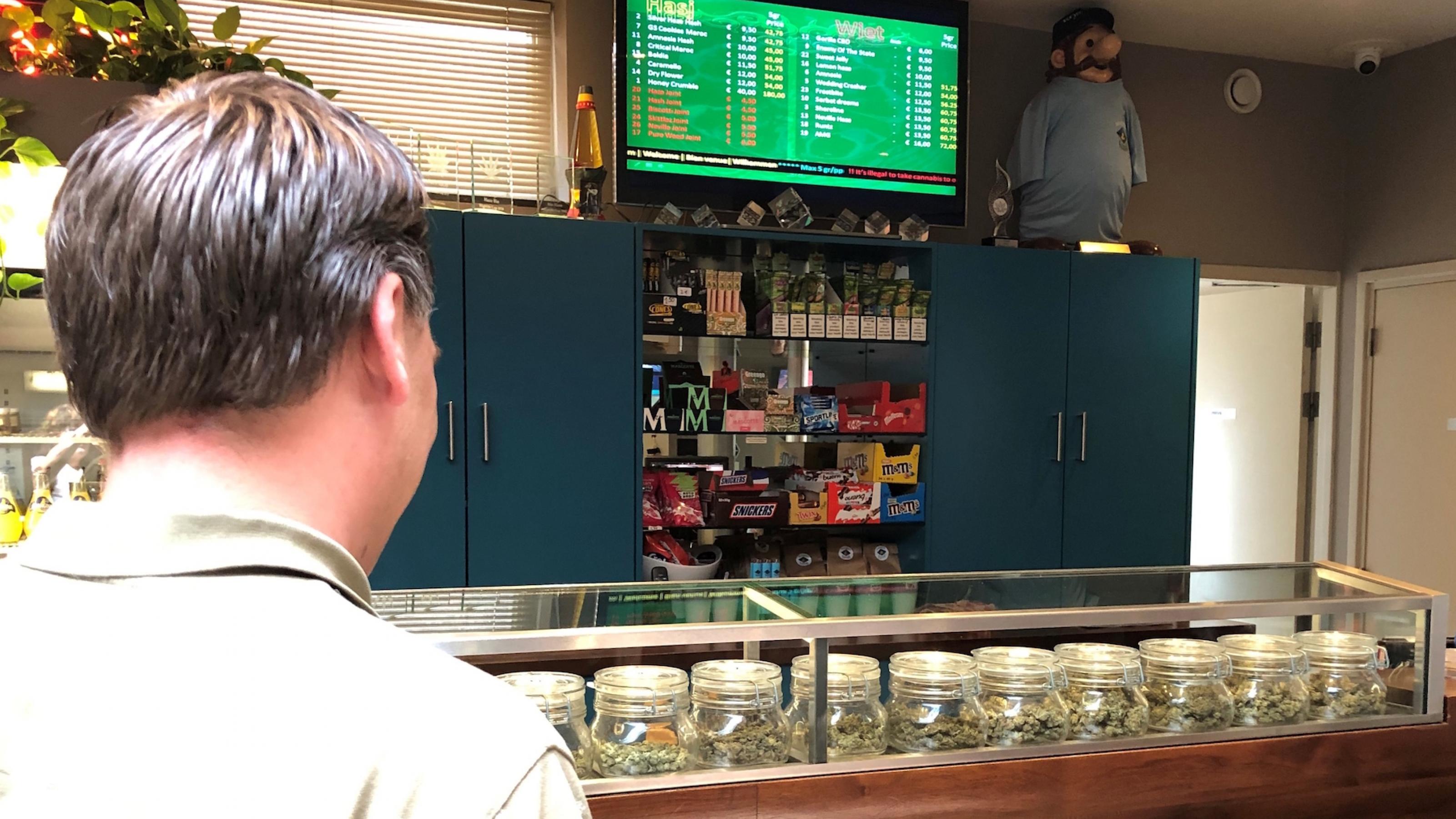 Ein Mann schaut in einem Coffeeshop auf eine Auswahl an verschiedenen Cannabis-Produkten hinter einem Tresen