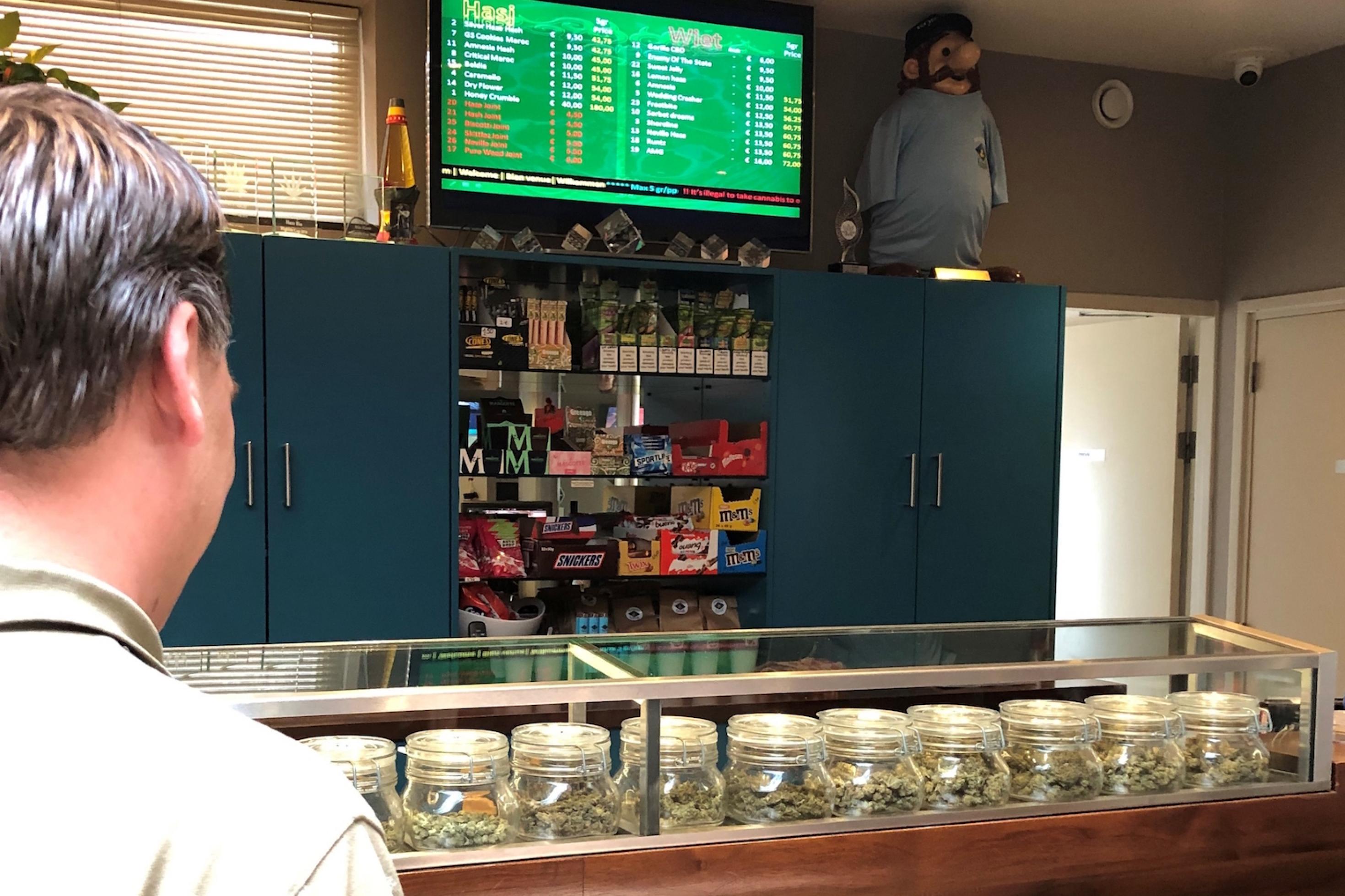 Ein Mann schaut in einem Coffeeshop auf eine Auswahl an verschiedenen Cannabis-Produkten hinter einem Tresen