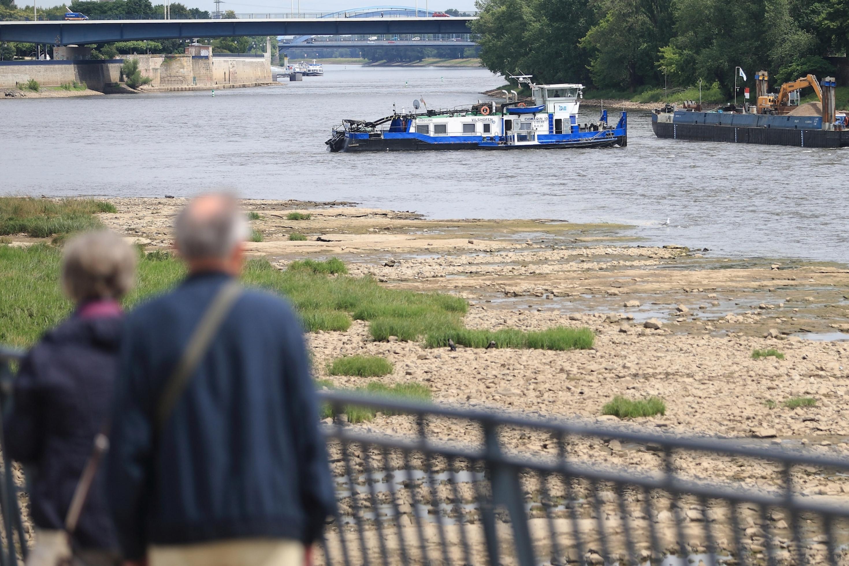 Zwei Menschen blicken auf den Strom. Unter ihnen liegt ein trockengefallener Felsbereich, im Hintergrund sind ein Schubschiff und eine Schute mit einem Bagger zu erkennen.