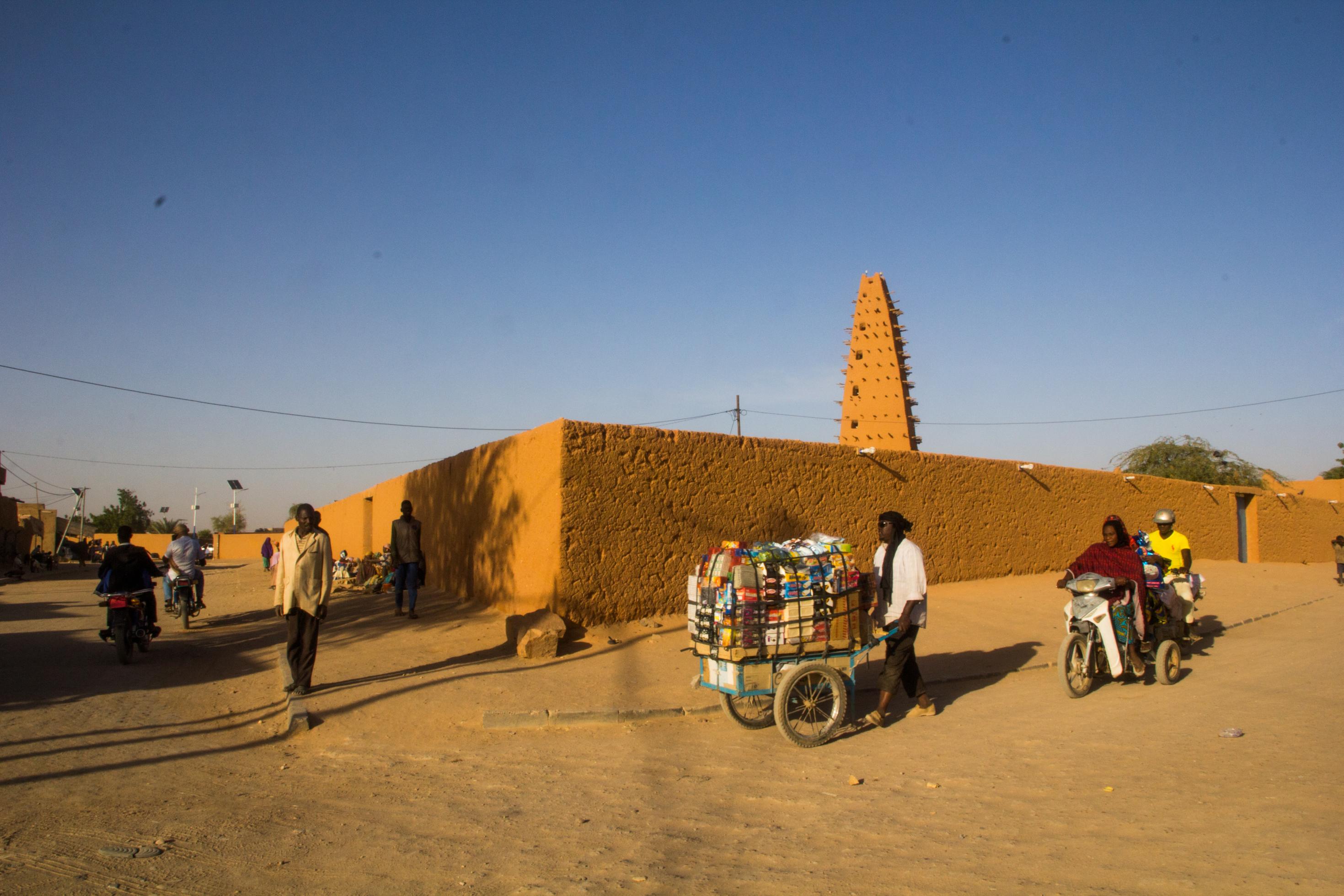 Der Lehmbau einer Moschee, im Vordergrund ein Mann mit Handkarren, darauf unterschiedliche Waren. Die Straße ist ungepflastert.
