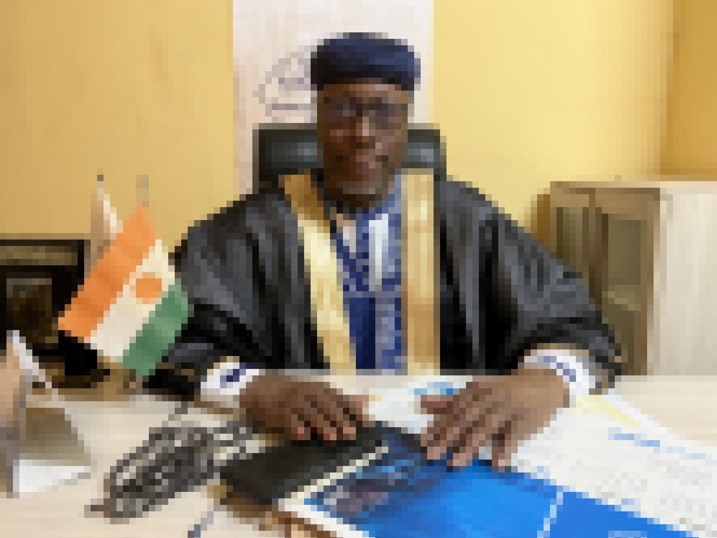 Hinter einem Schreibtisch sitzt ein mittelalter Mann, vor sich auf dem Tische eine Flagge von Niger und eine Gebetskette. Er trägt ein blaues, traditionelles Gewand mit breiter, goldener Borte. t flachem, blauen