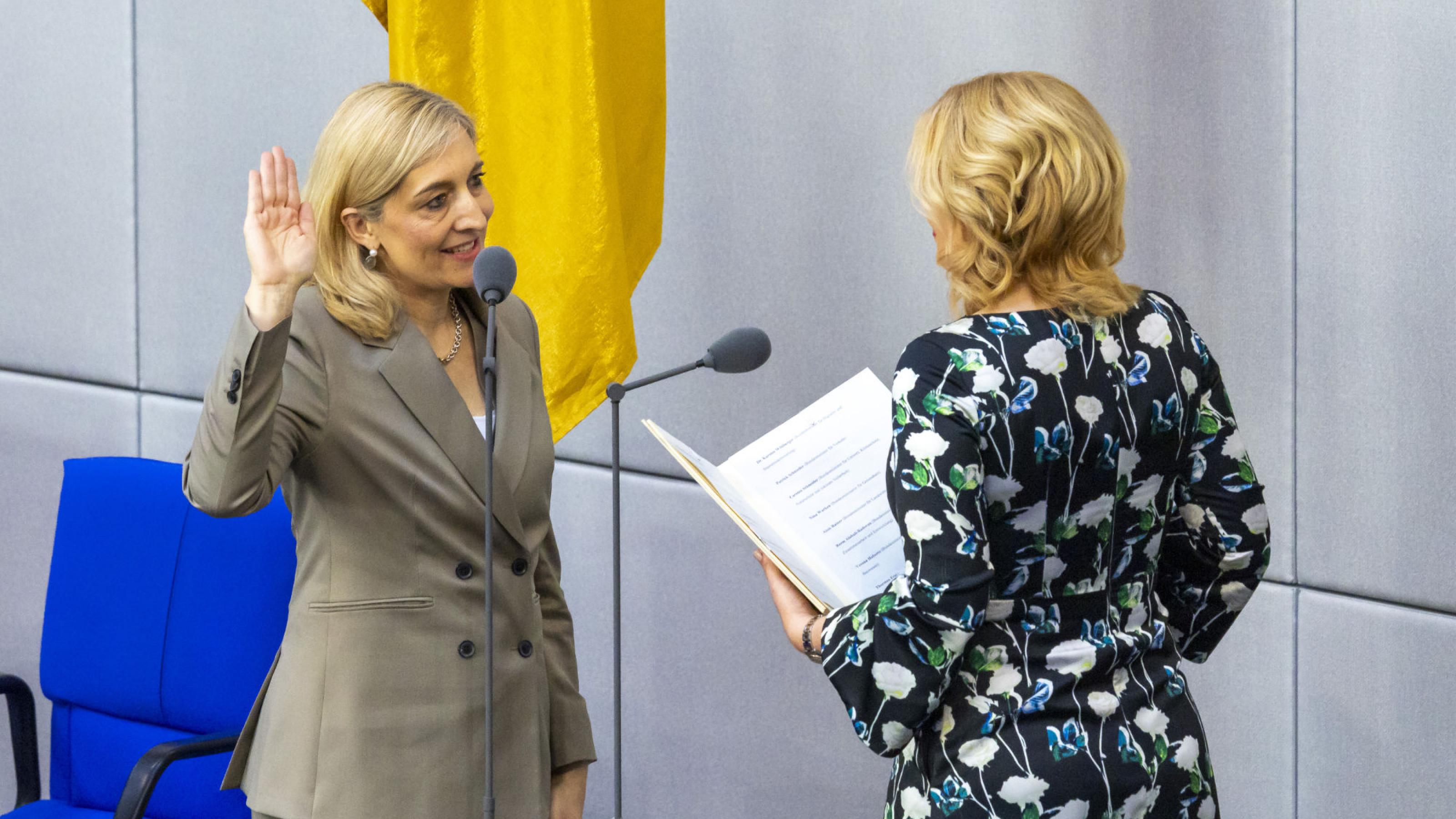 Vor einer Deutschlandflagge im Bundestag leistet Nina Warken mit erhobener Hand ihren Amtseid, von hinten zu sehen ist Bundestagspräsidentin Julia Klöckner mit einer Kladde.