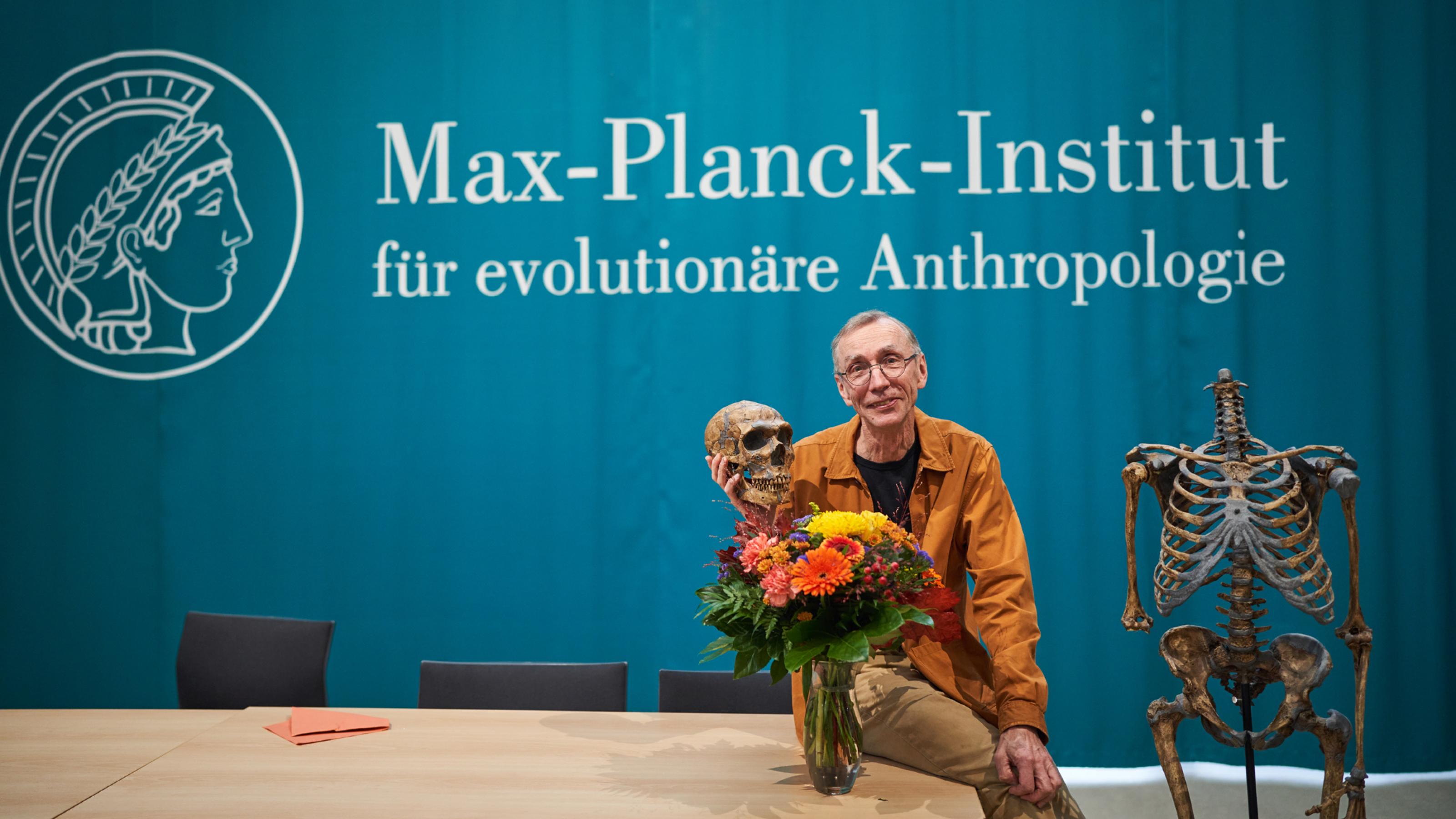 Ein blauer Hintergrund mit der Aufschrift „Max-Plank-Institut für evolutionäre Anthropologie“. Davor sitzt auf einem beigen Holztisch ein Mann in bräunlich-oranger Jacke und beiger Hose, einen großen Blumenstrauß in einer Vase vor sich und in der rechten Hand den Schädel eines Neandertalers tragend. Rechts von ihm steht das kopflose, restliche Skelett des Neandertalers.