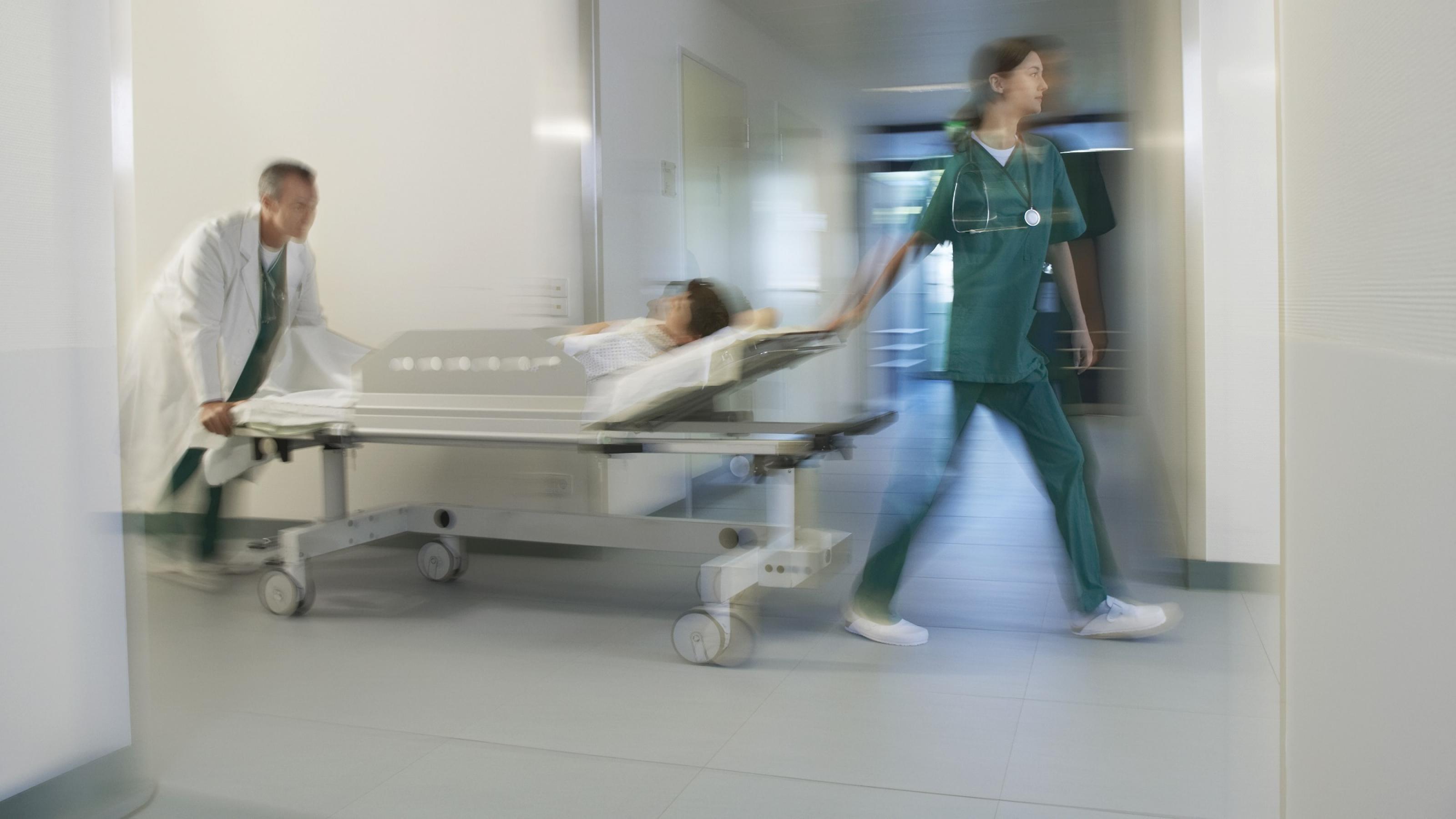 Ein Krankenpfleger schiebt einen Notfall-Patienten durch einen Krankenhausflur. Eine Pflegerein lenkt vorne. Sie haben es eilig.