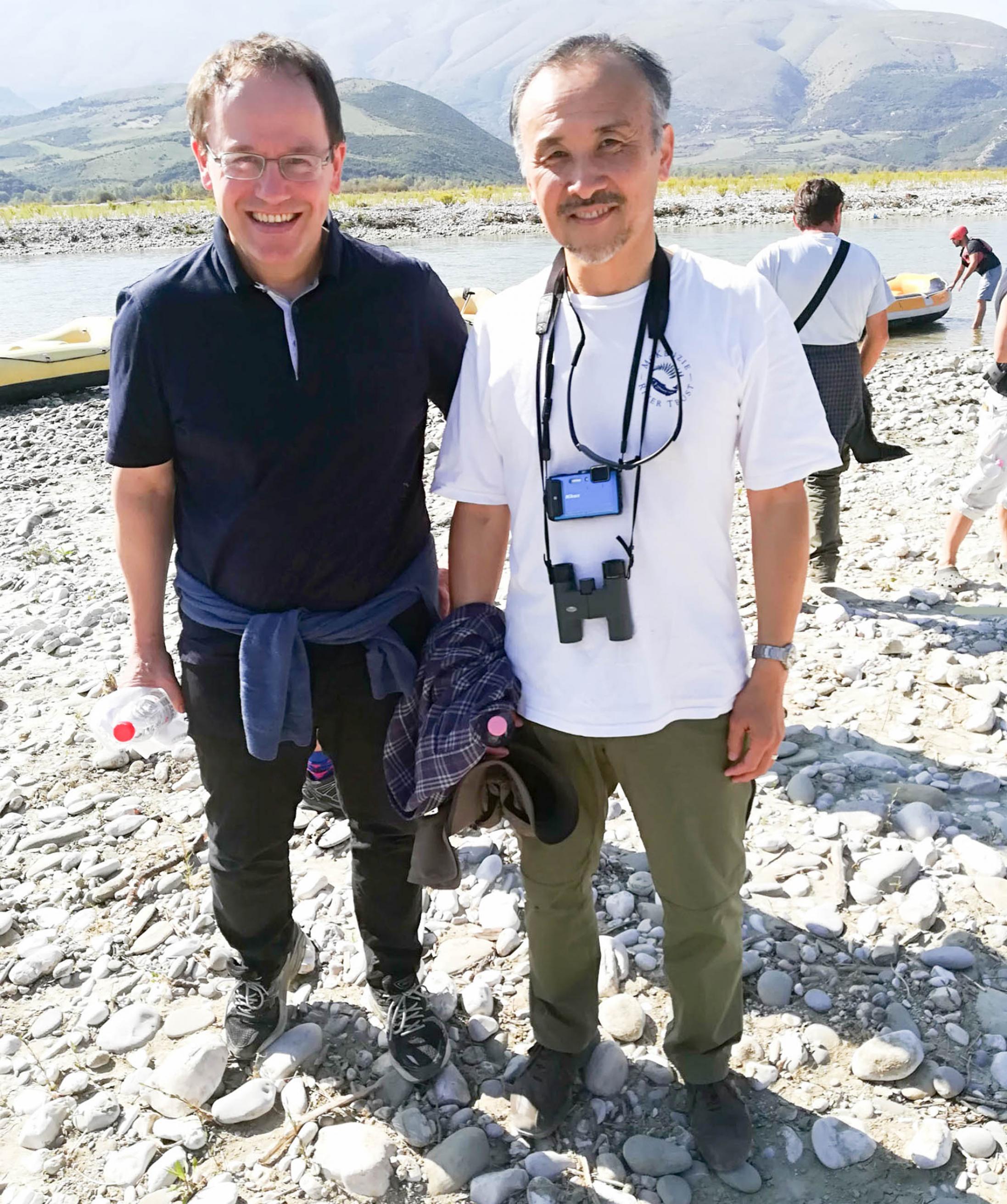 Zwei Forscher stehen auf Schotter an einem Fluss und schauen in die Kamera. Der rechte Mann hat einen Fotoapparat und ein Fernglas vor der Brust hängen.