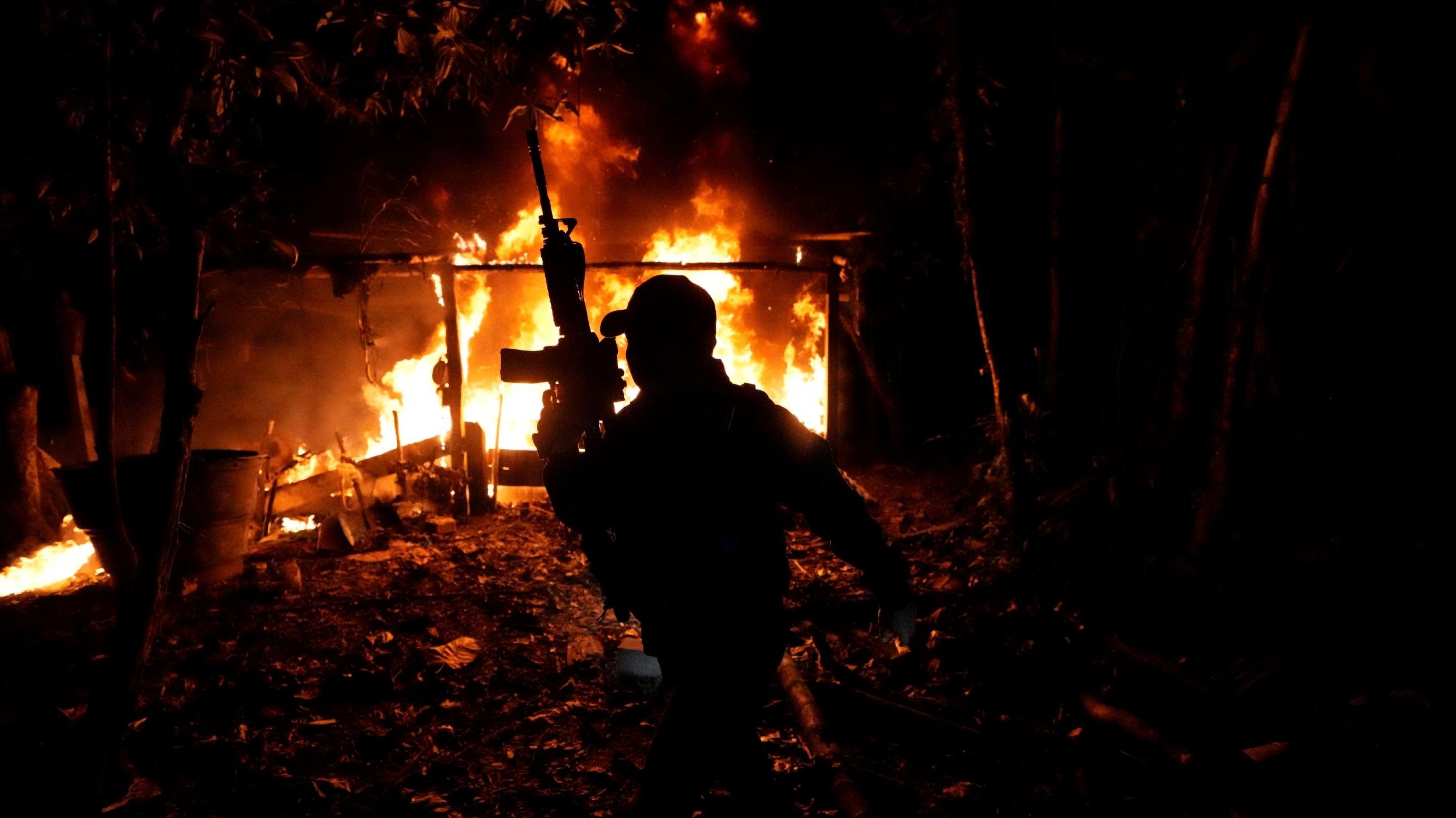 Vor dem Hintergrund eines im Dunkeln brennenden Hauses sieht man die Silhouette eines Mannes, der ein Maschinengewehr nach oben hält