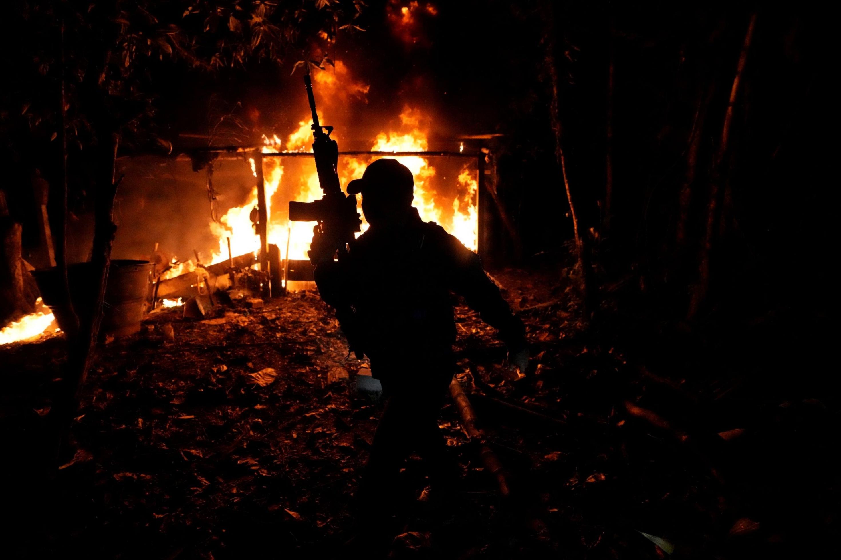 Vor dem Hintergrund eines im Dunkeln brennenden Hauses sieht man die Silhouette eines Mannes, der ein Maschinengewehr nach oben hält