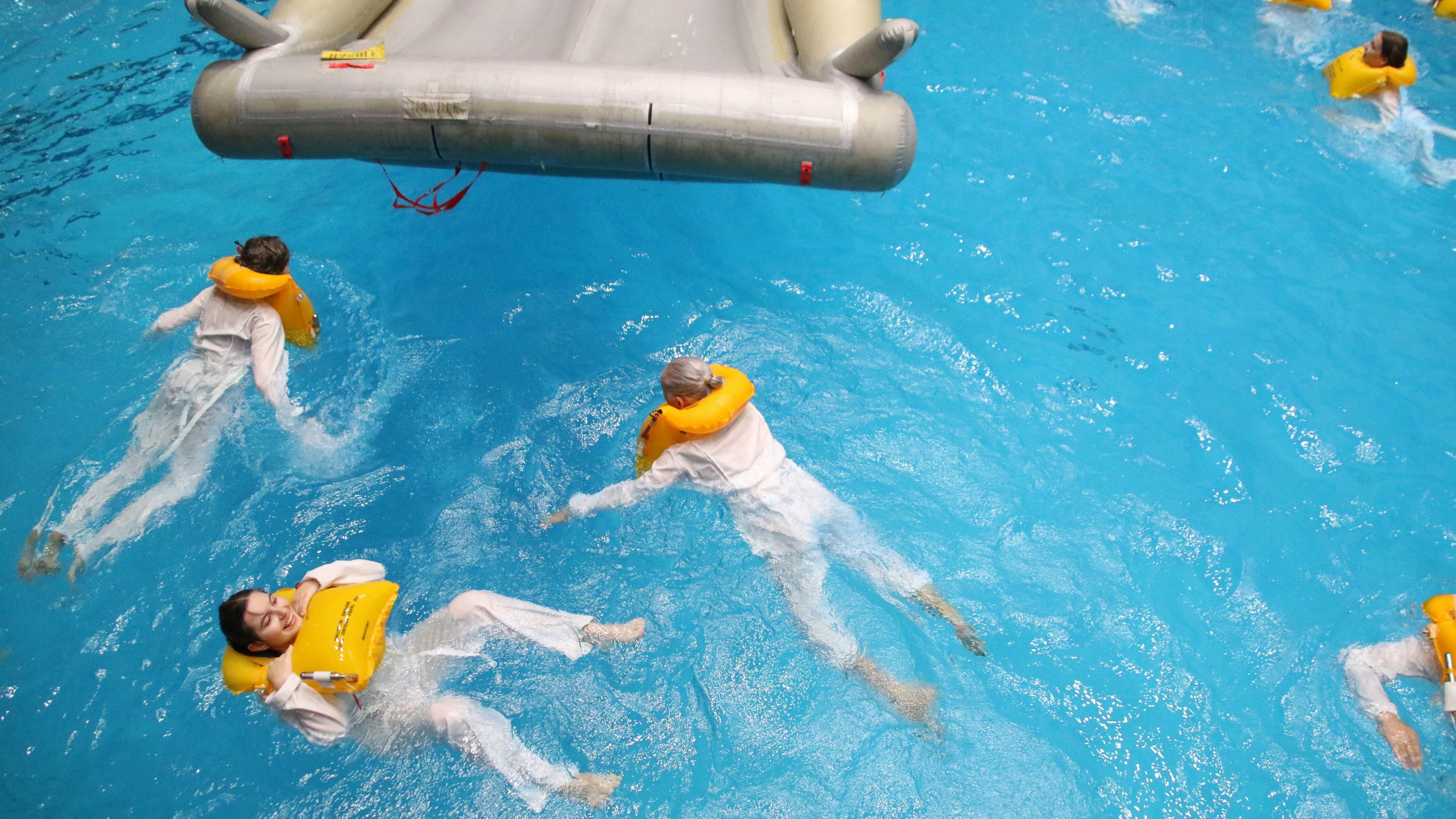 Flugbegleiterinnen schwimmen in einem Hallenbad neben einem Rettungsfloß