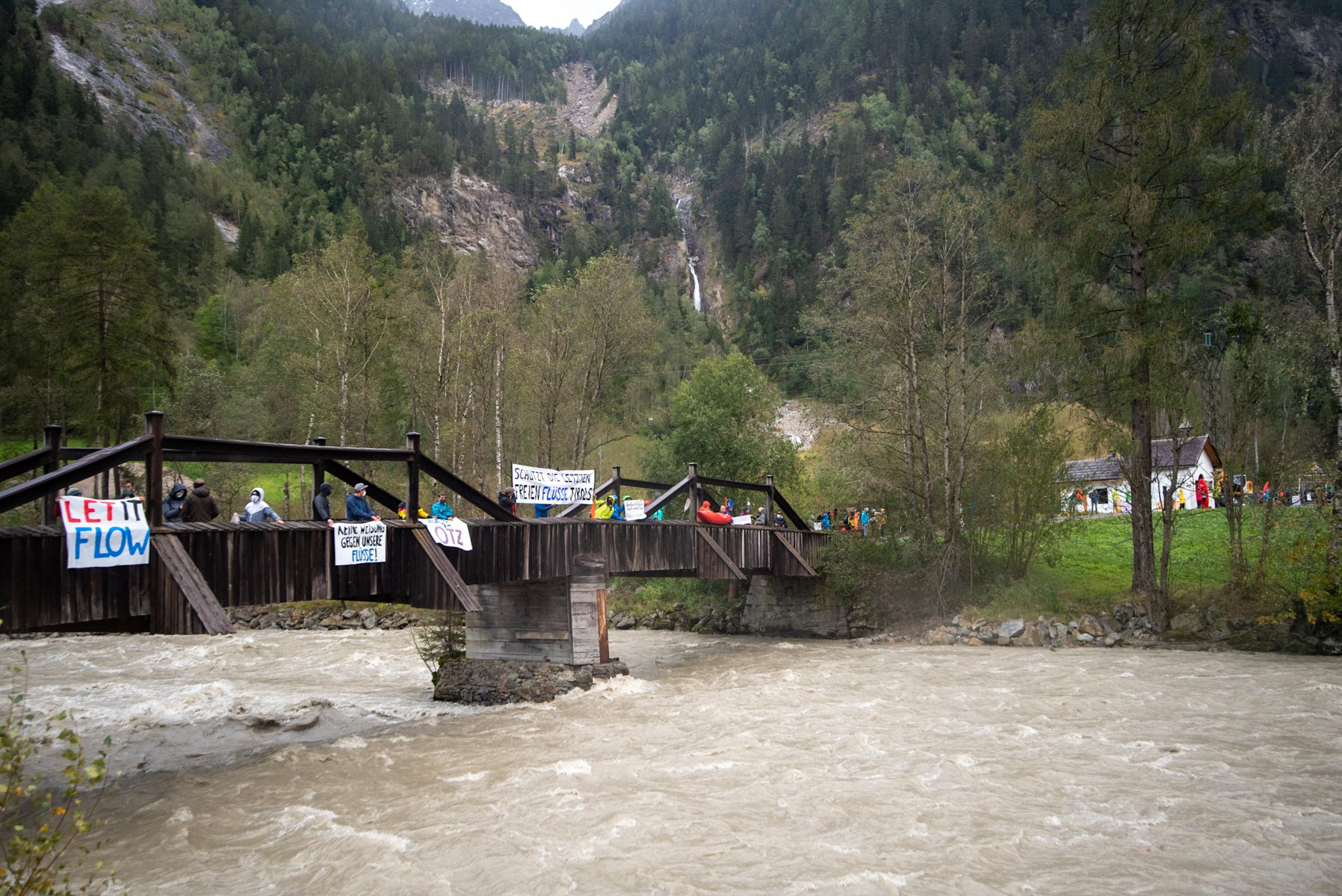 Ein Berg mit einem herunter stürzenden Bach im Hintergrund, vorne eine Holzbrücke über die Ache mit protestierenden Menschen.
