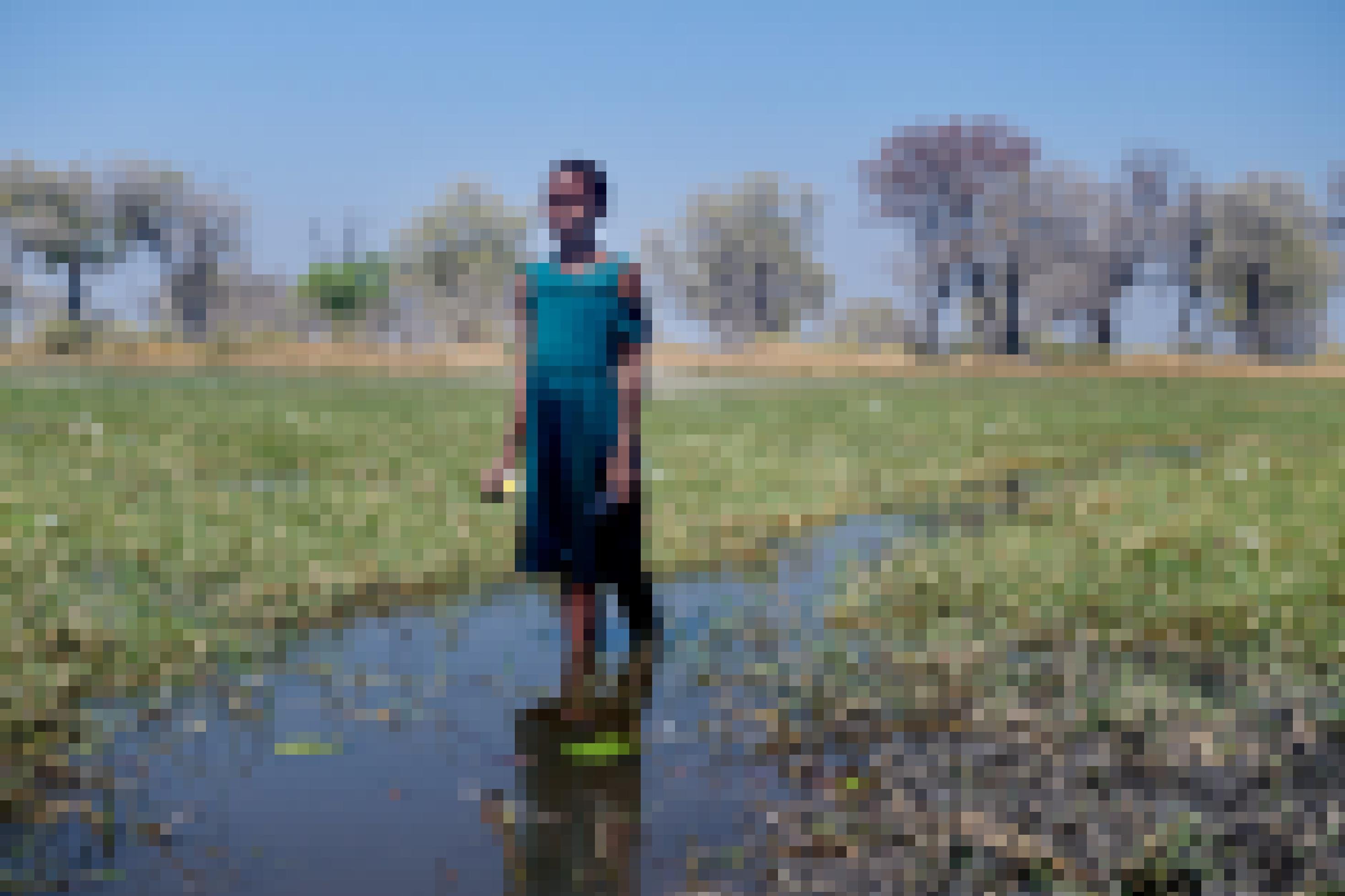 Das Mädchen steht bis zu den Knöcheln im Wasser in der weiten Landschaft, in der Hand hält sie eine Wasserlilie, im Hintergrund sind Bäume zu sehen.