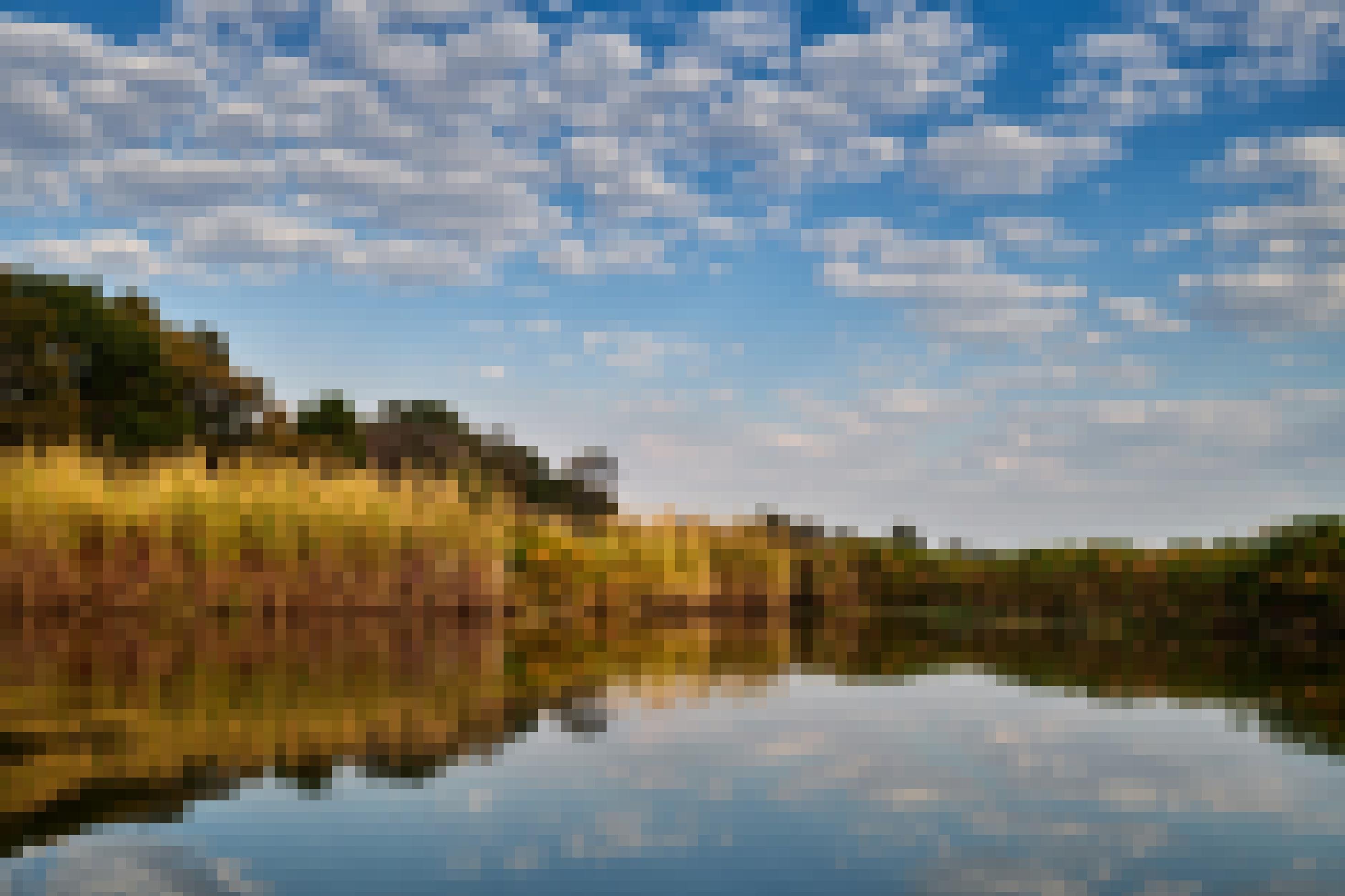 Im Fluss spiegeln sich die Wolken des sonst blauen Himmels, die Ufer sind mit Schilf bewachsen