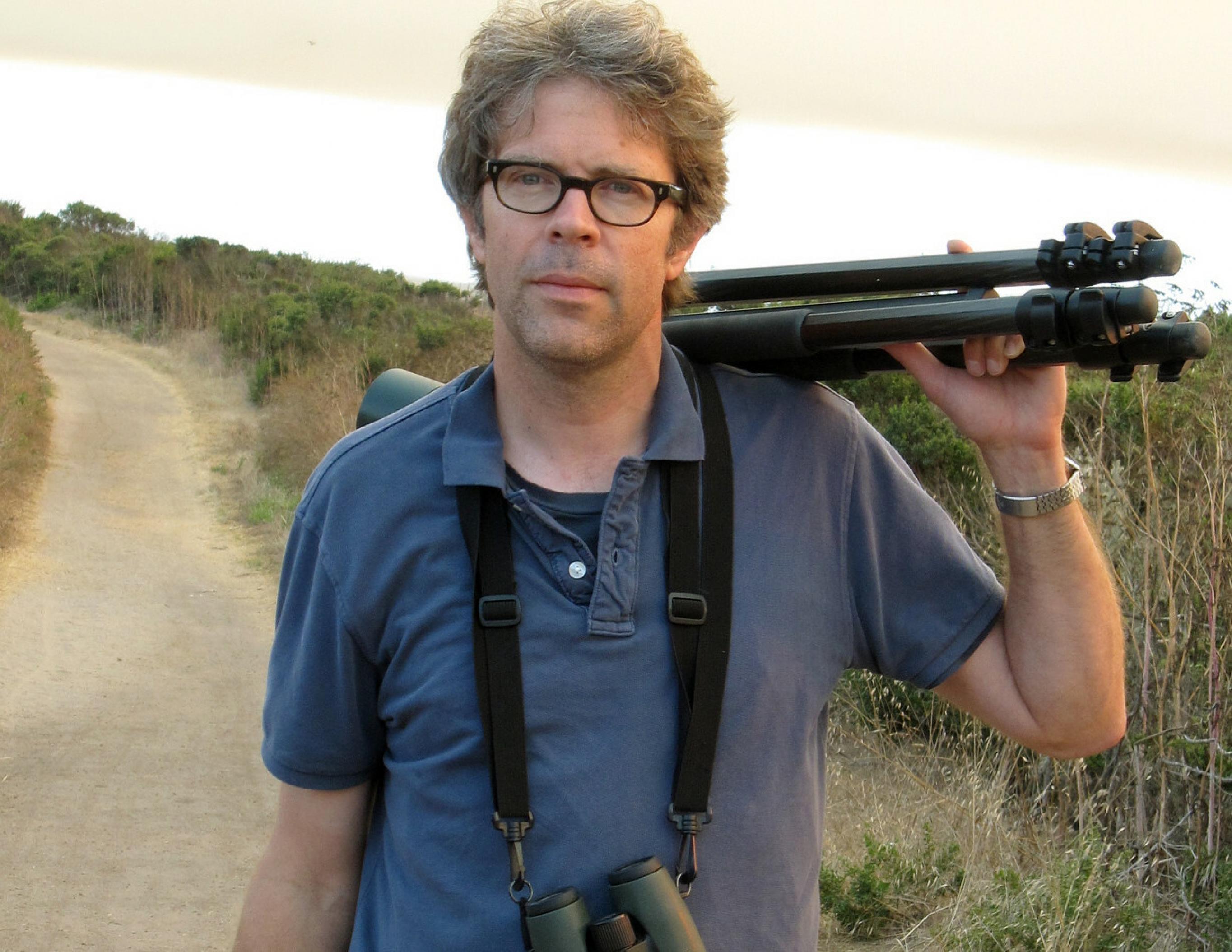 Jonathan Franzen mit Stativ auf der Schulter und einem Fernglas umgehängt in einer Hügellandschaft stehend.