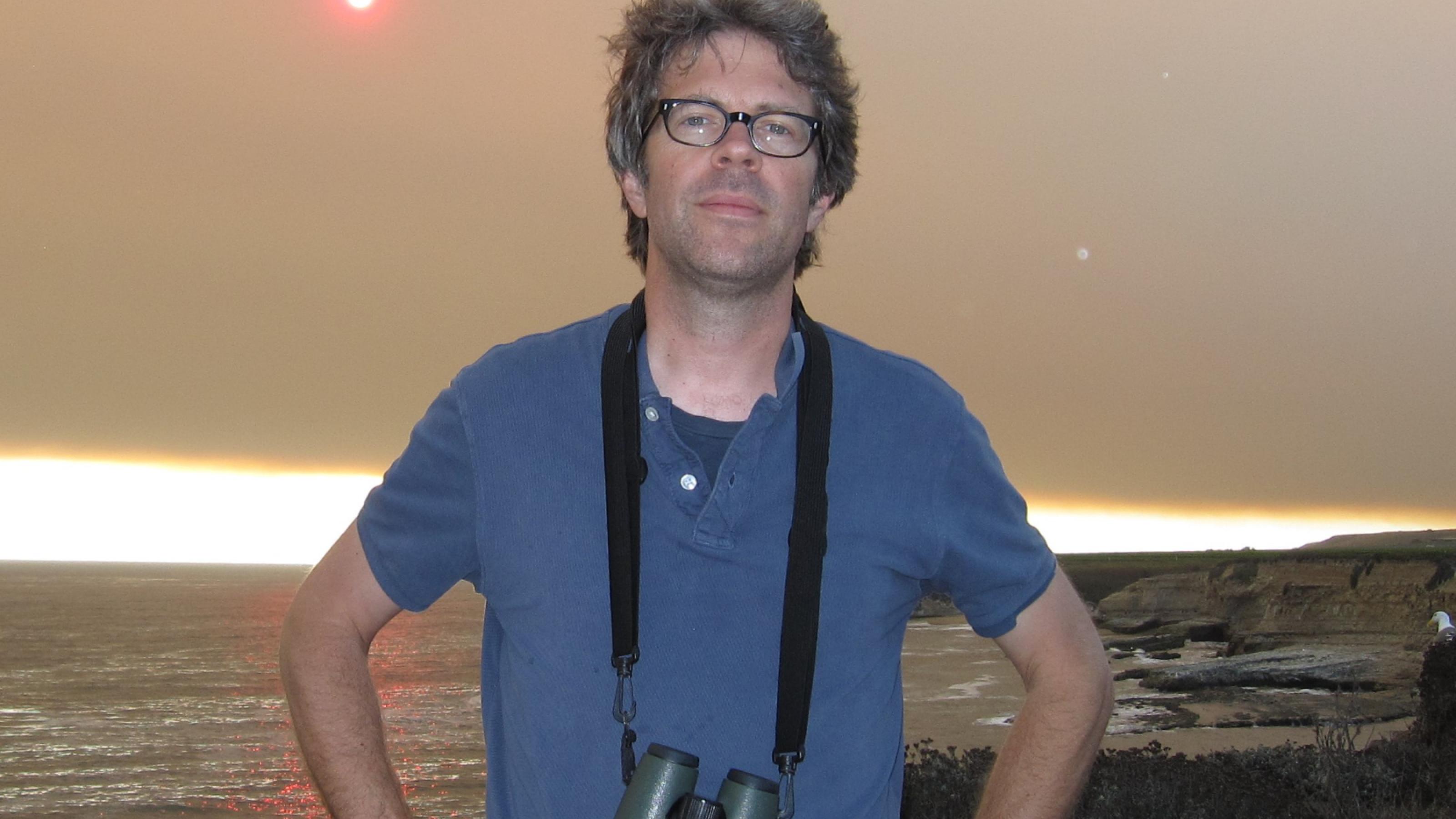 Jonathan Franzen steht mit einem Fernglas um den Hals im T-Shirt an einer Meeresküste.