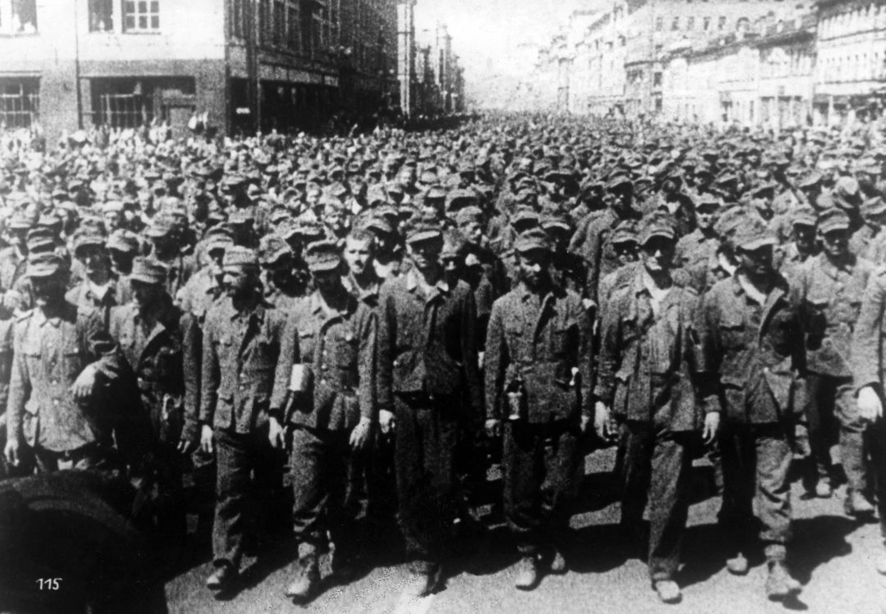 Ein altes Foto in schwarz-weiß zeigt eine endlose Menge an deutschen Soldaten ohne Waffen, die auf einer breiten Straße zwischen Häusern laufen.