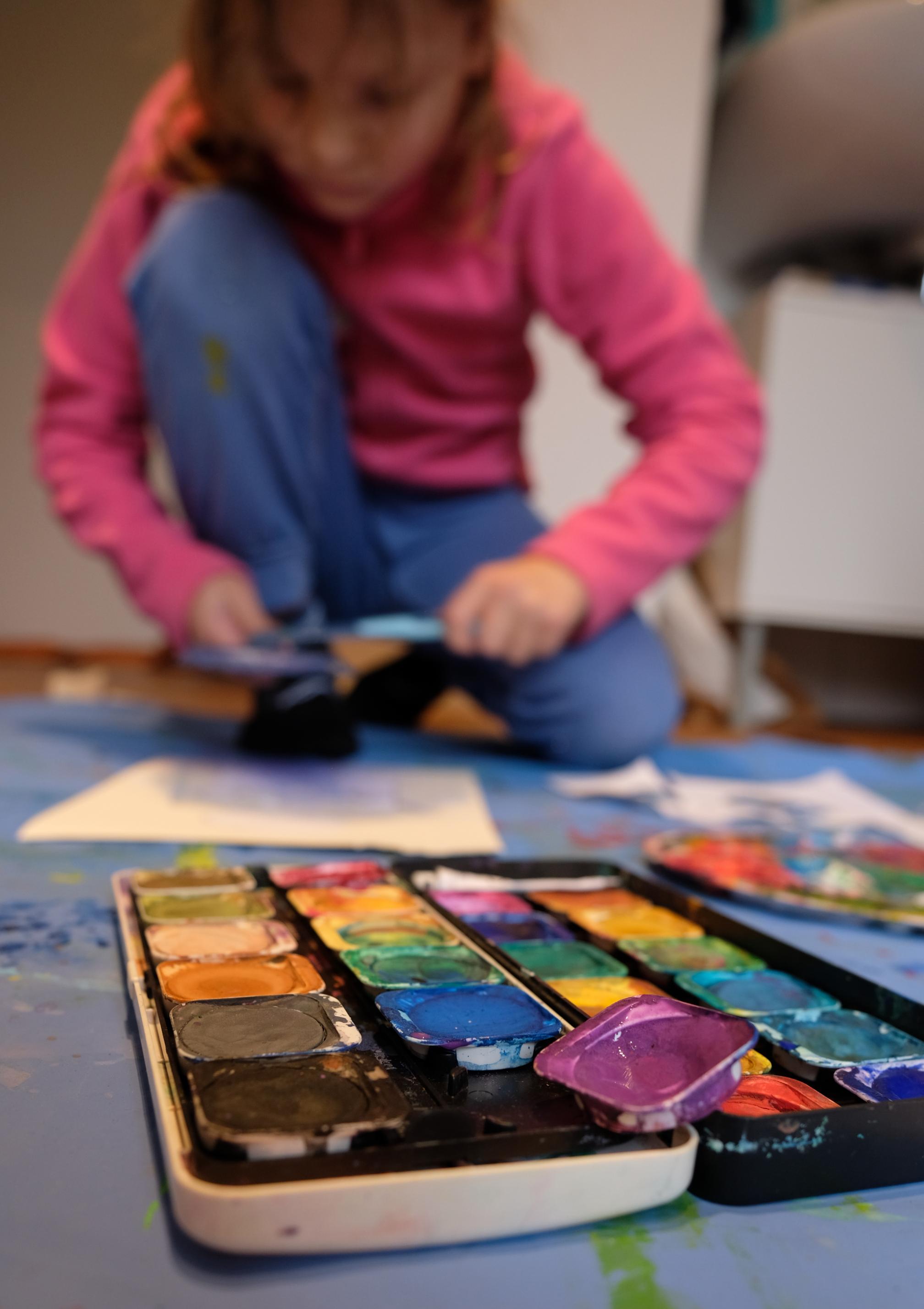Im Hintergrund des Bildes hockt die achtjährige Orla und sprüht mit Farben über Papier. Im Vordergrund sieht man ganz groß den wild ausgebeuteten Malkasten. Alle Farben wurden schon oft benutzt.