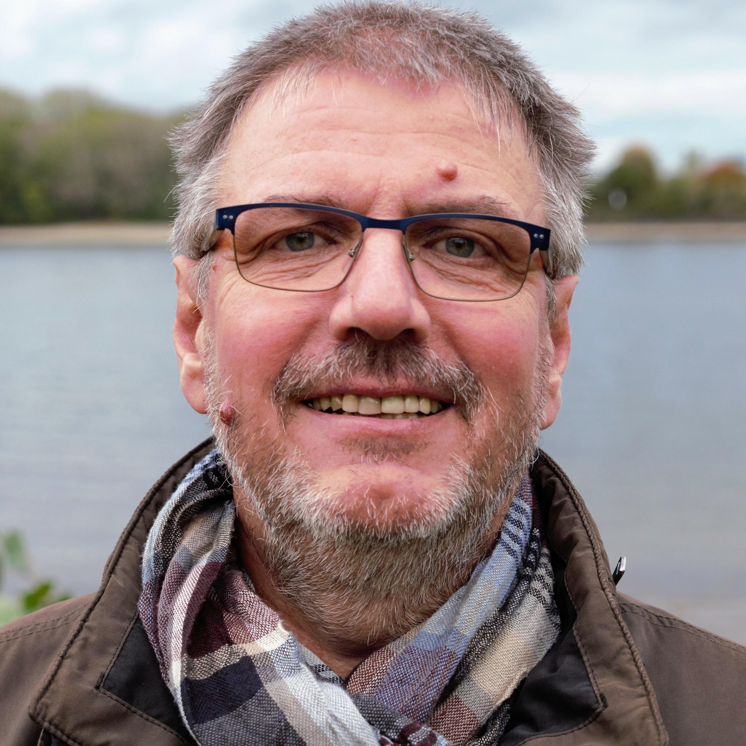 Grauhaariger Mann mit Brille, Schal und brauner Jacke blickt zum Betrachter, im unscharfen Hintergrund fließt der Fluss Rhein unter grau bewölkten Himmel.