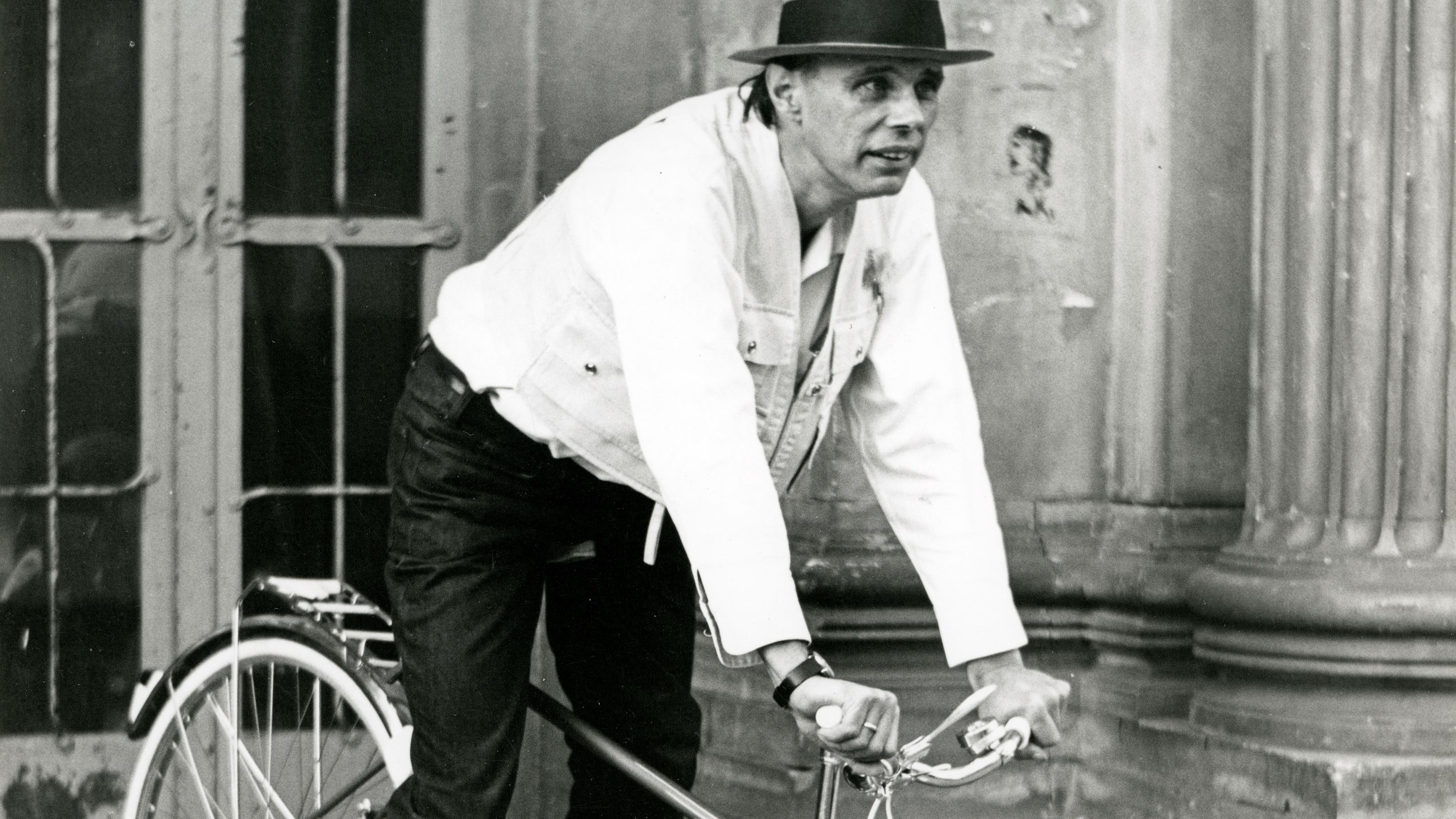 Beuys fährt ii Mountainbike-Stil, gekleidet mit weißem Hemd und dunkler Hose, drei flache Steinstufen hinunter.