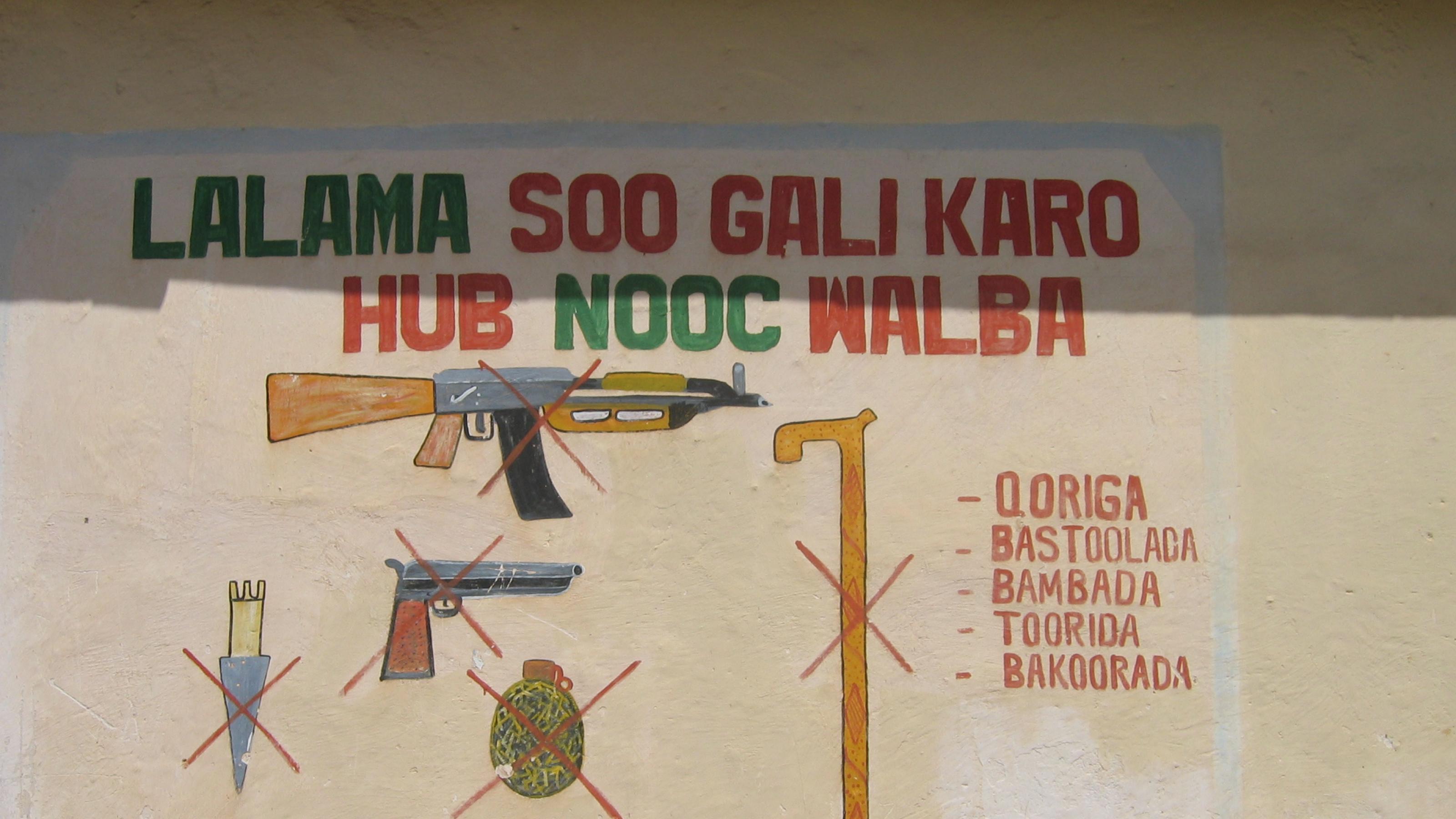 Ein gemaltes Hinweisschild auf der Außenmauer eines Gebäudes. Durchgestrichen sind verschiedene Waffen, also eine Kalashnikow, ein Messer, eine Handgranate und ein Gehstock.