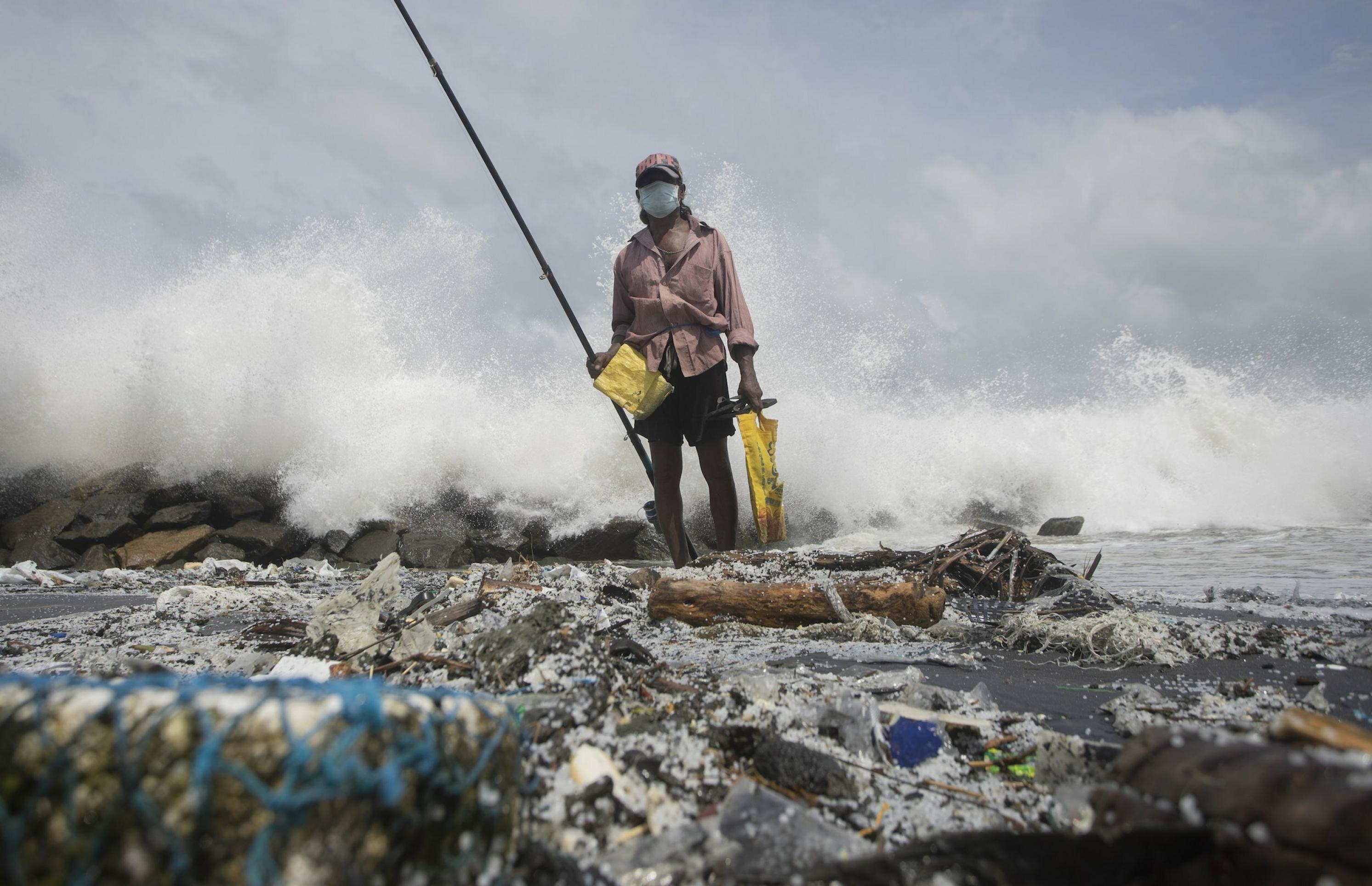 Fischer inmitten von Müll am Strand mit einer langen Angel in der Hand.