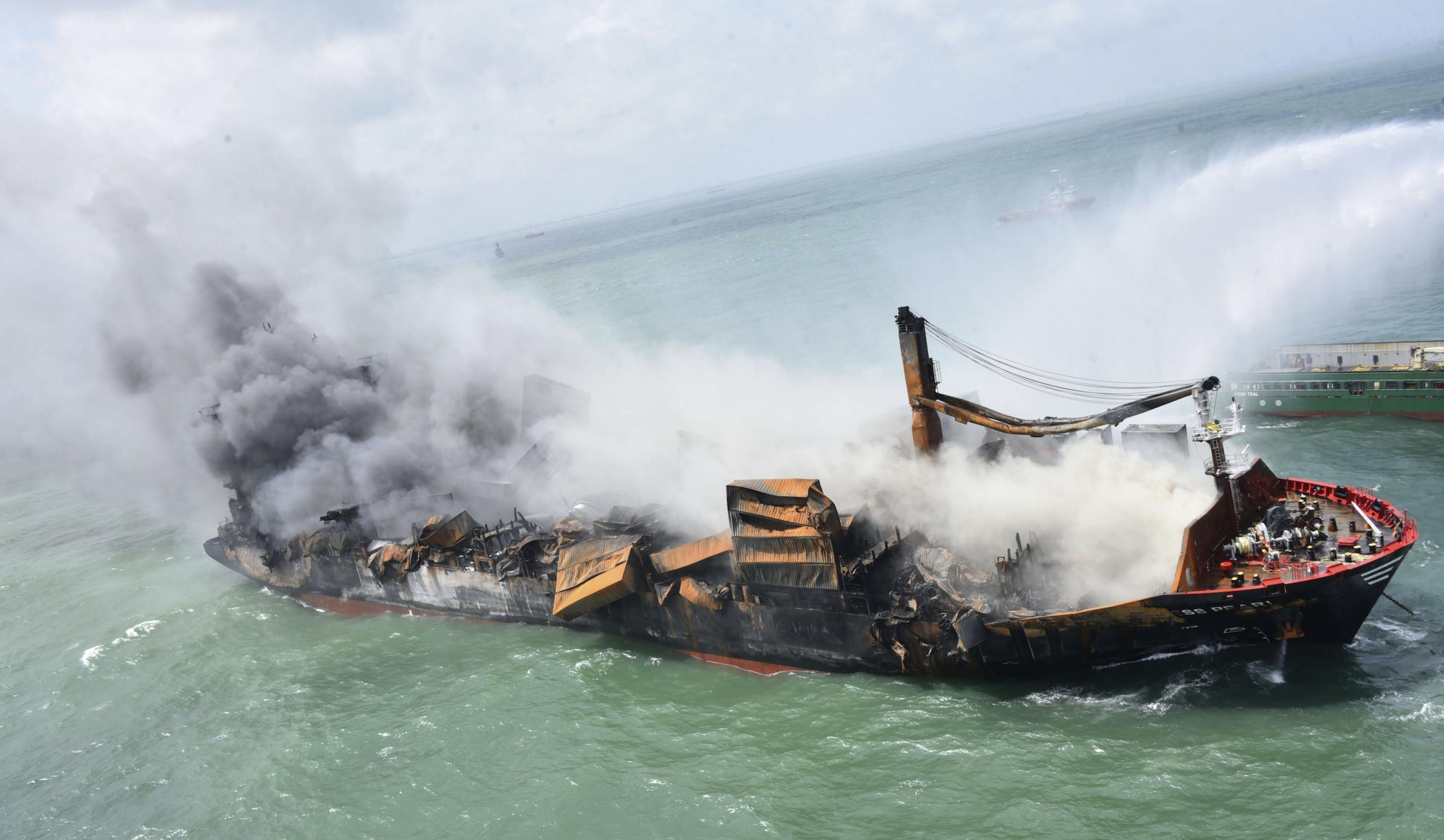 Ein sinkender, brennender Frachter, von rechts Wasser von einem Löschschiff.
