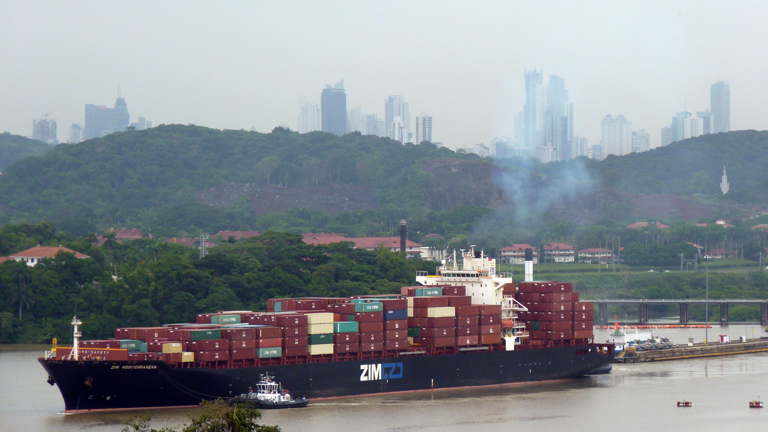 Ein schwer beladenes Containerschiff, im Hintergrund die Hochhaus-Skyline von Panama-Stadt.