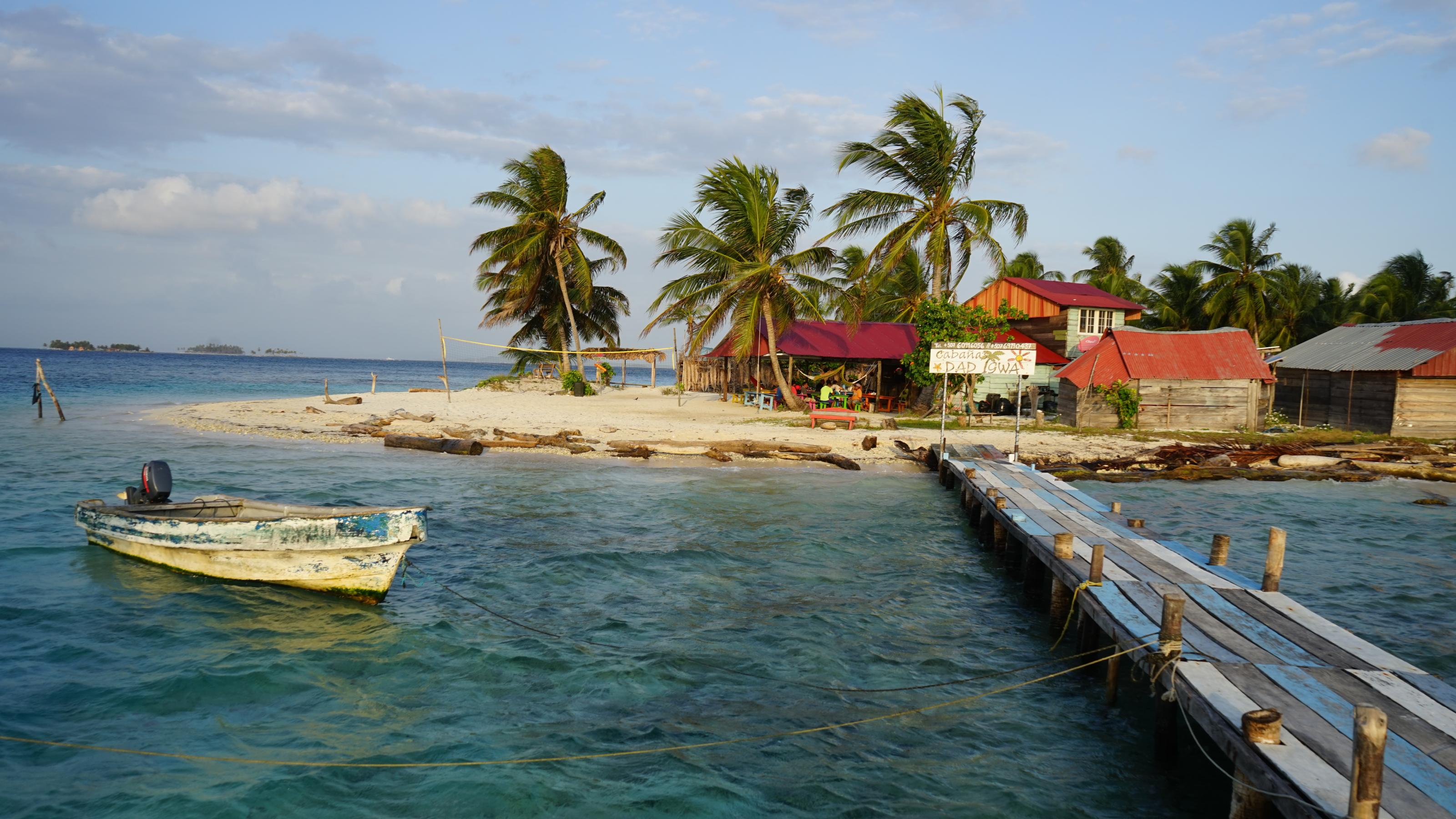 Ein Atoll auf Gunayala, mit Holzsteg und einem vor Anker liegenden Boot, im Hintergrund bunte Hütten.