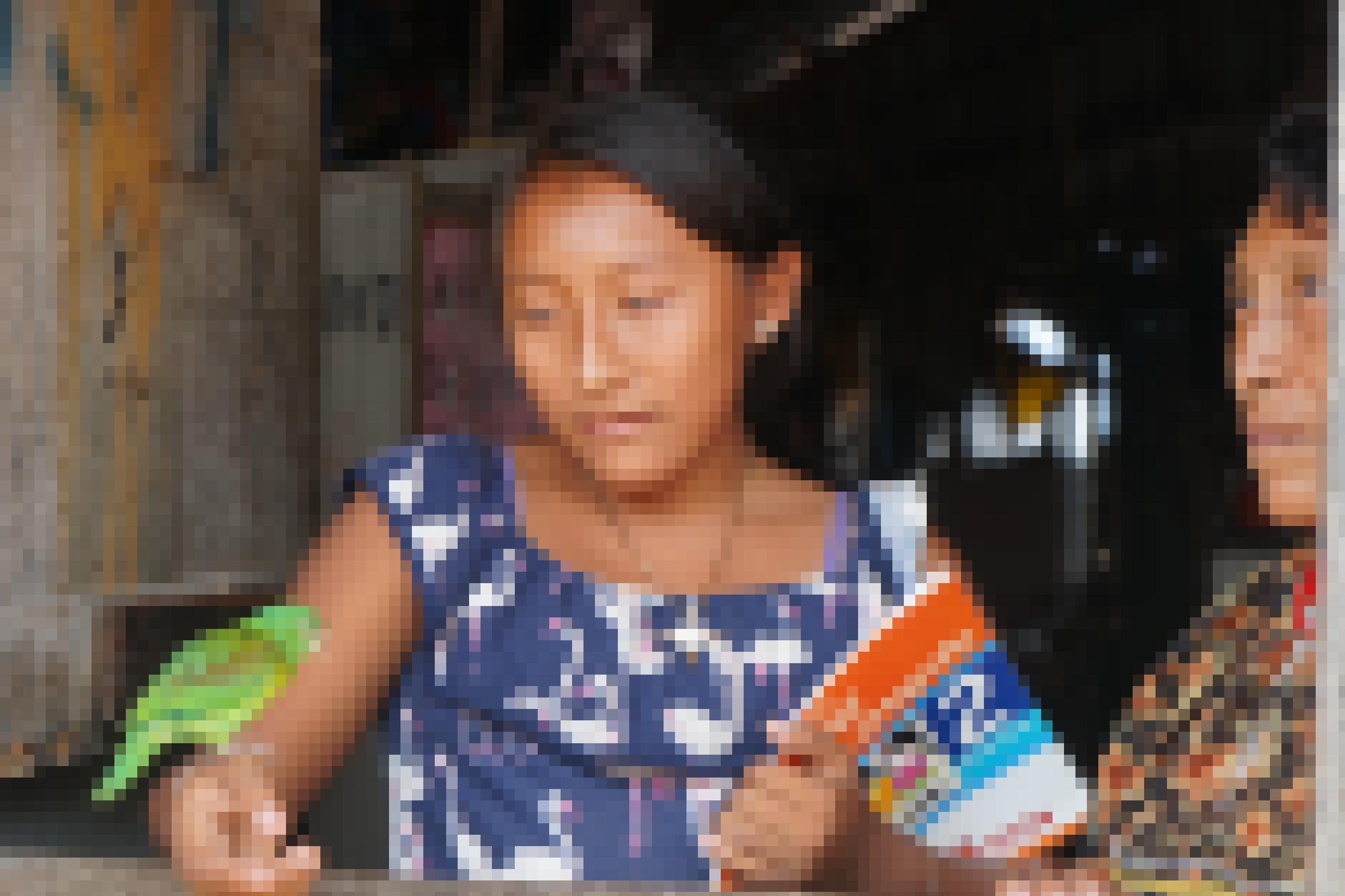 Ein Kuna-Mädchen sitzt an einem Holzfenster, in der linken Hand ein Schulbuch, mit der rechten spielt es mit einem grünen Papagei.