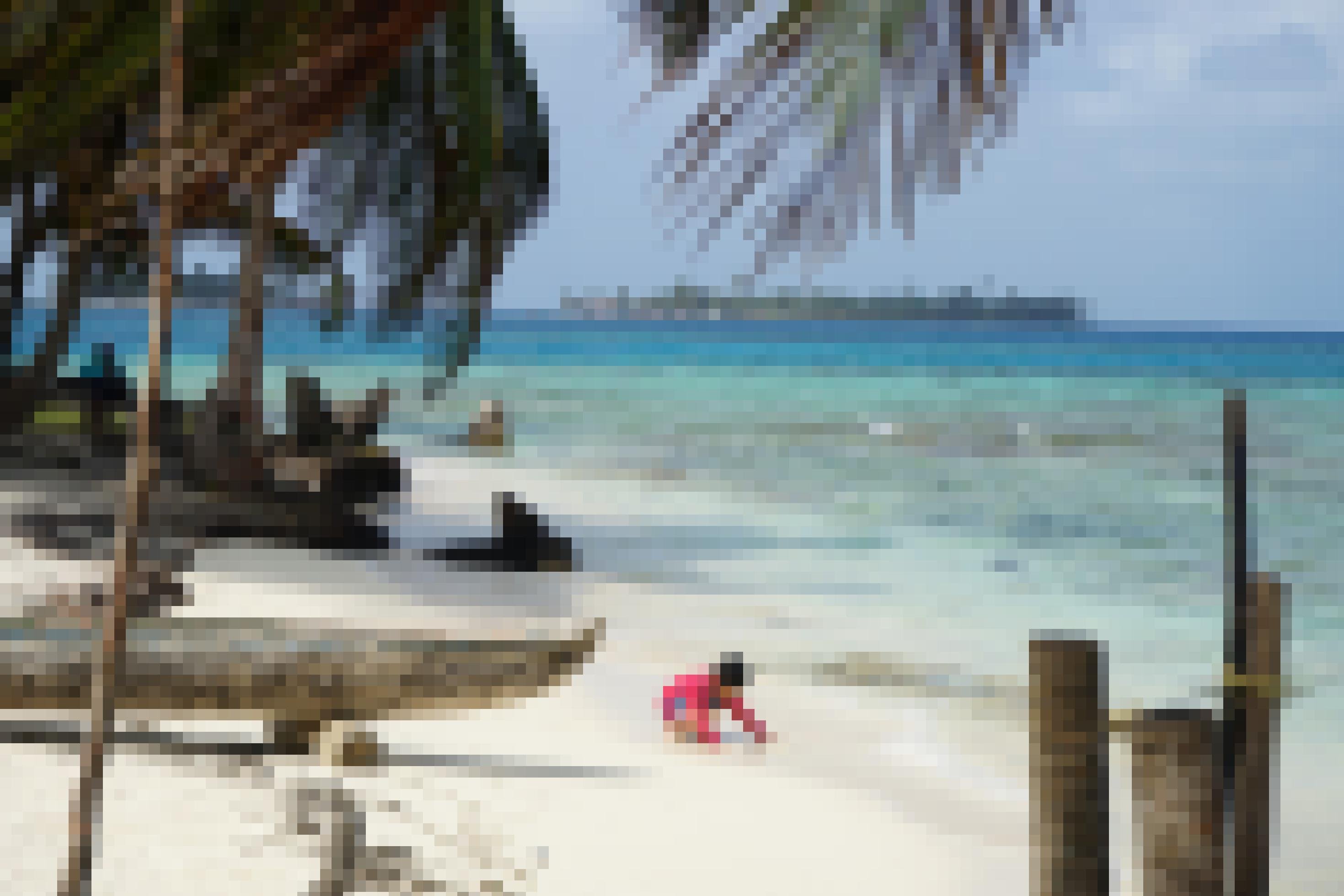 Ein heller Korallen-Sandstrand mit einem Holzboot, davor spielend ein kleiner Junge, dahinter türkisfarbenes Meer.