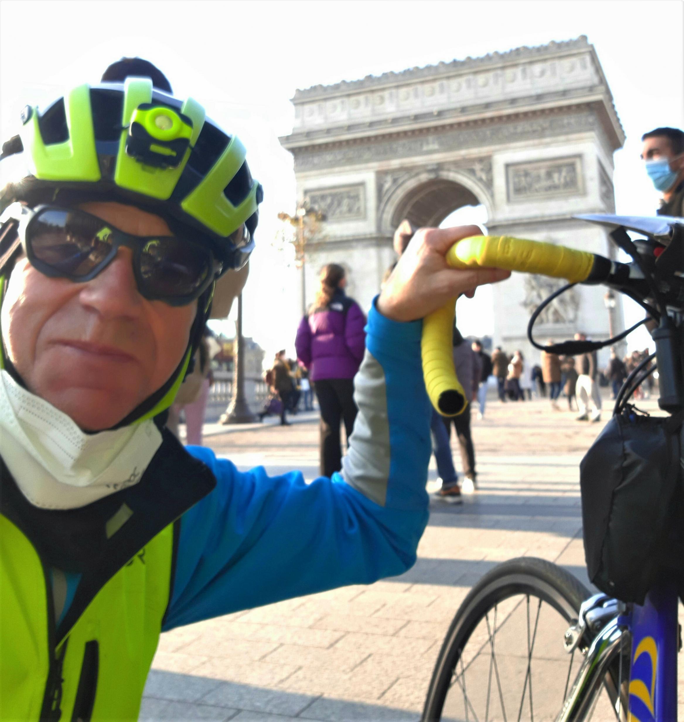 Im Vordergrund des Pariser Triumpf-Bogens schießt der behelmte und bebrillte Reporter mit der Rechten ein Selfie, während der mit der Linken den Lenker seines Rennrads hält.