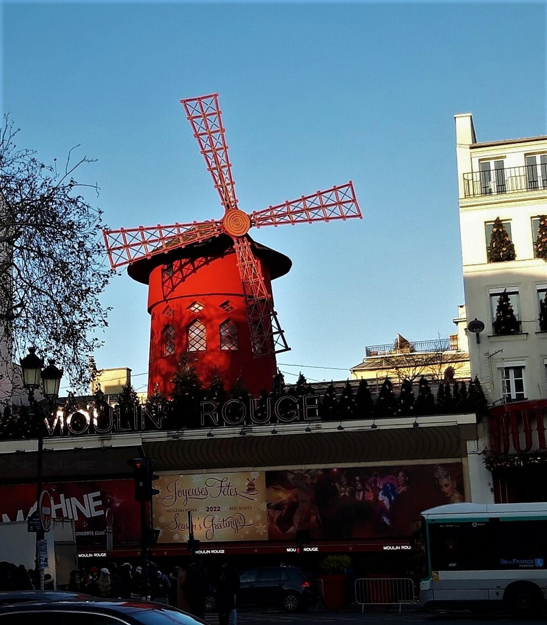 Vor pulsierendem Straßenverkehr ragt auf einem Flachbau eine in leuchtendem Rot gestrichene Windmühle auf.