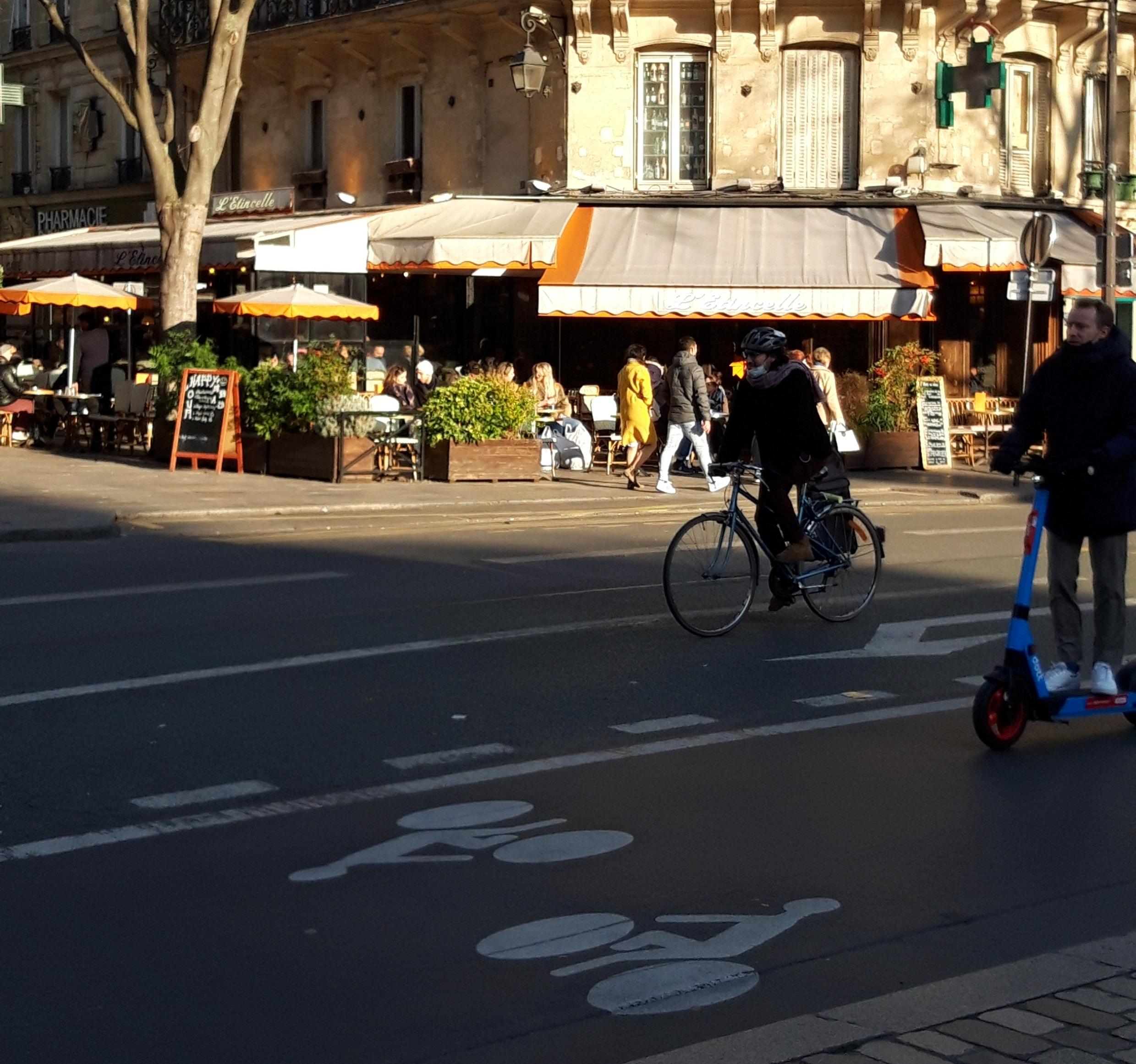 Auf einer breiten Rad-Spur fahren ein Fahrrad und ein E-Roller ins Bild, dessen Hintergrund von einer sonnenbeschienenen Terrasse eines Restaurants bestimmt wird.
