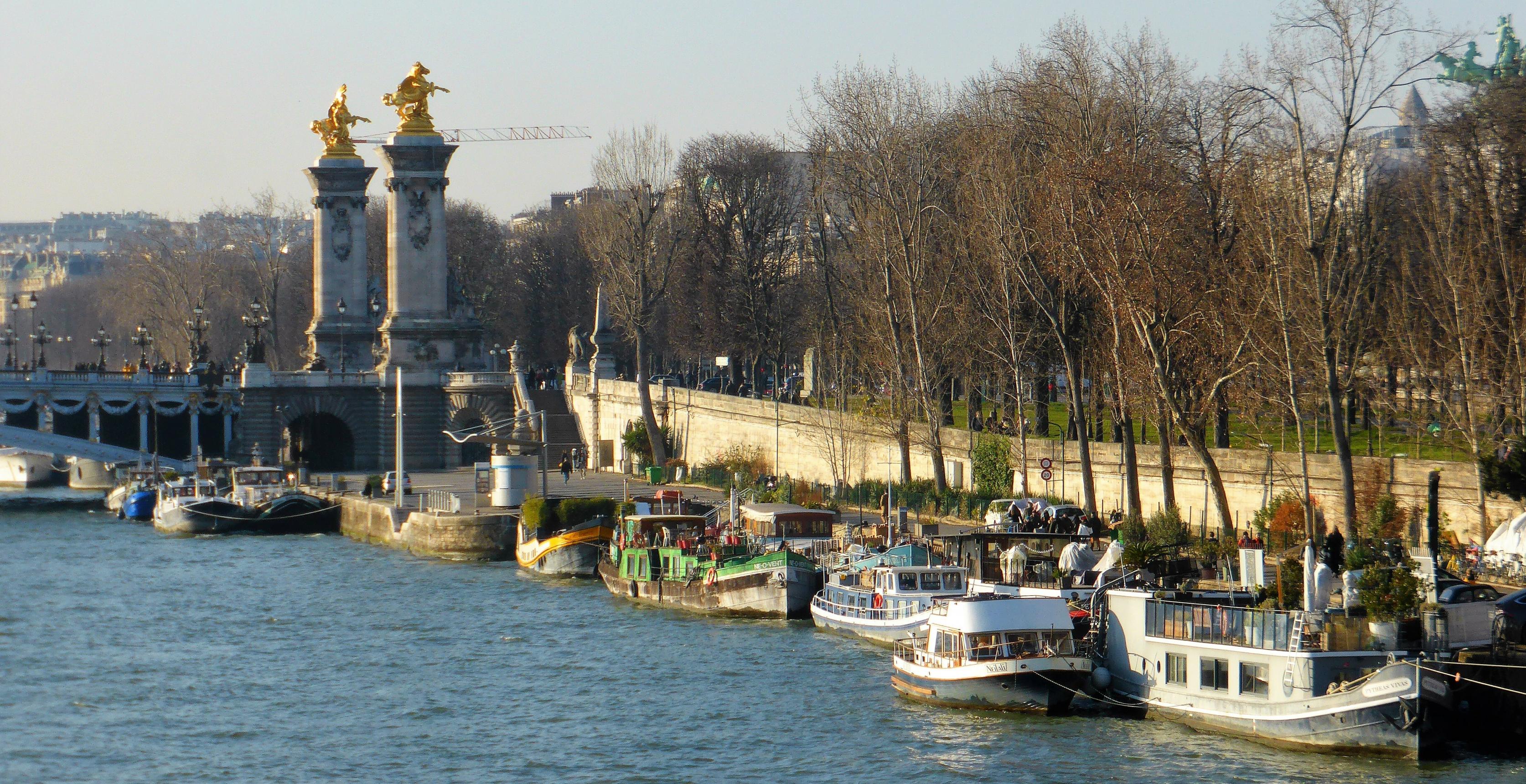 Vor dem neobarocken Brückenkopf oberhalb des Seine-Wassers dümpeln knapp ein Dutzend Boote am von der Sonnen strahlend ausgeleuchteten Flussufer.