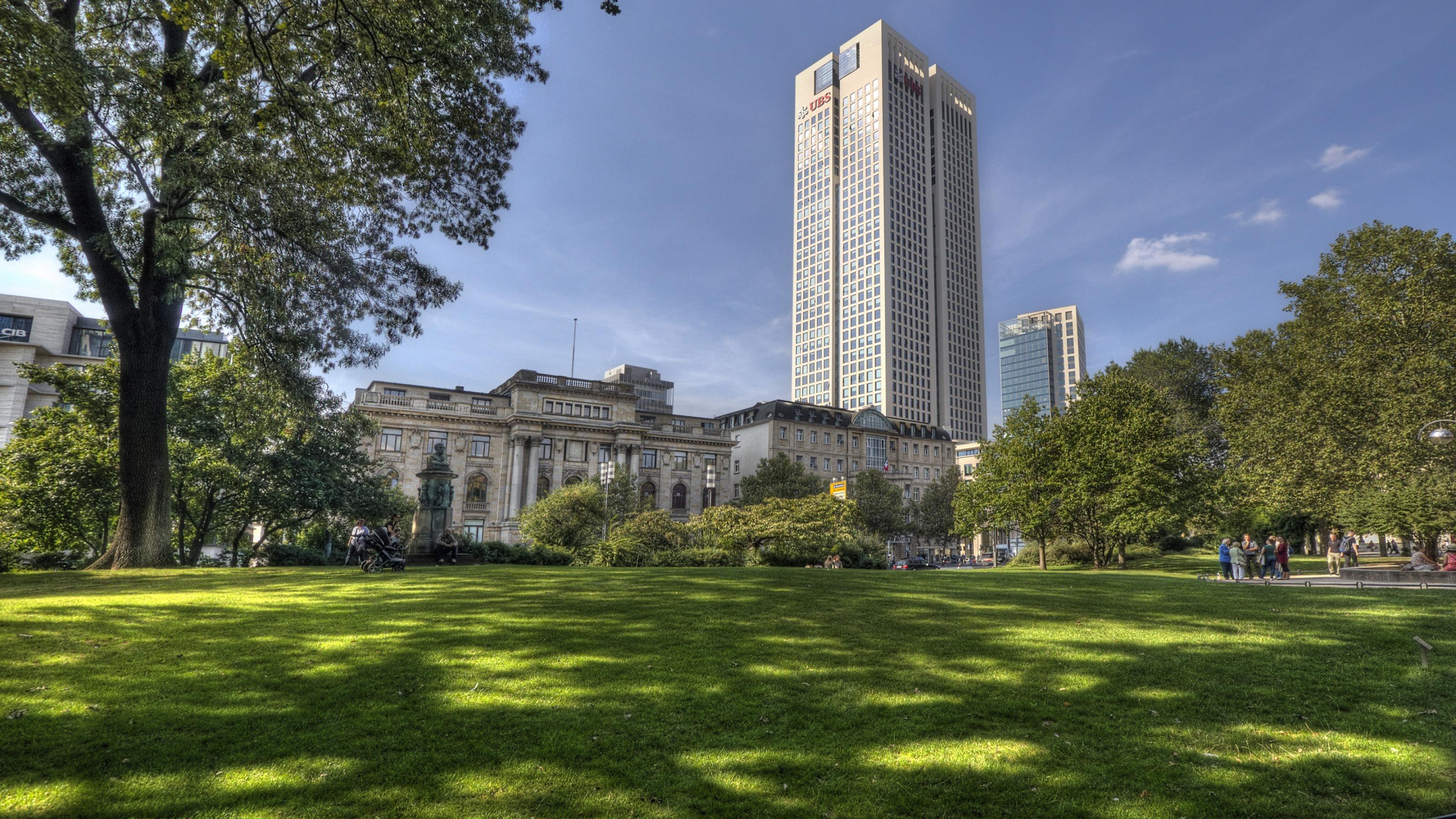 Im Vordergrund ein schöner Park mit grüner Wiese und Schatten spendendem Baum, im Hintergrund ein Hochhaus