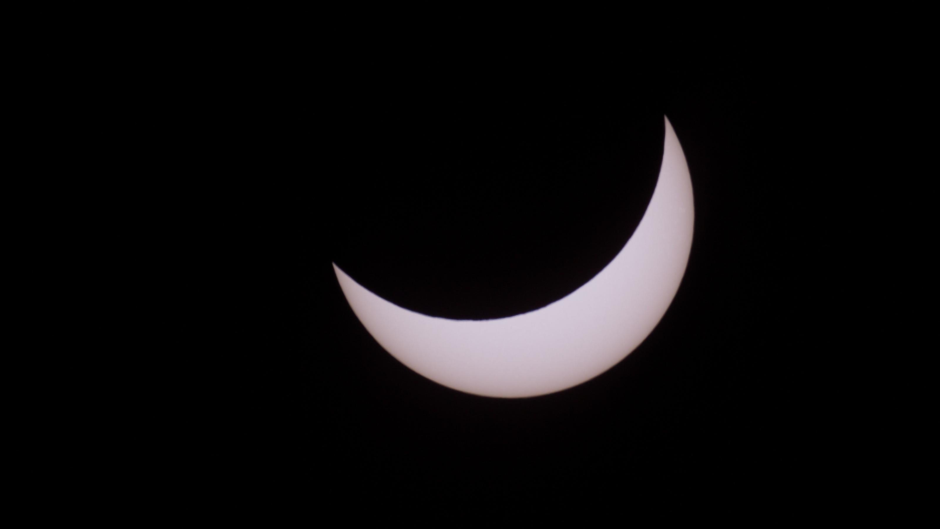 Auf dem Bild ist eine gelblich-weiße, sichelartige Sonne vor schwarzem Hintergrund zu sehen, da sie teilweise vom Mond bedeckt ist.
