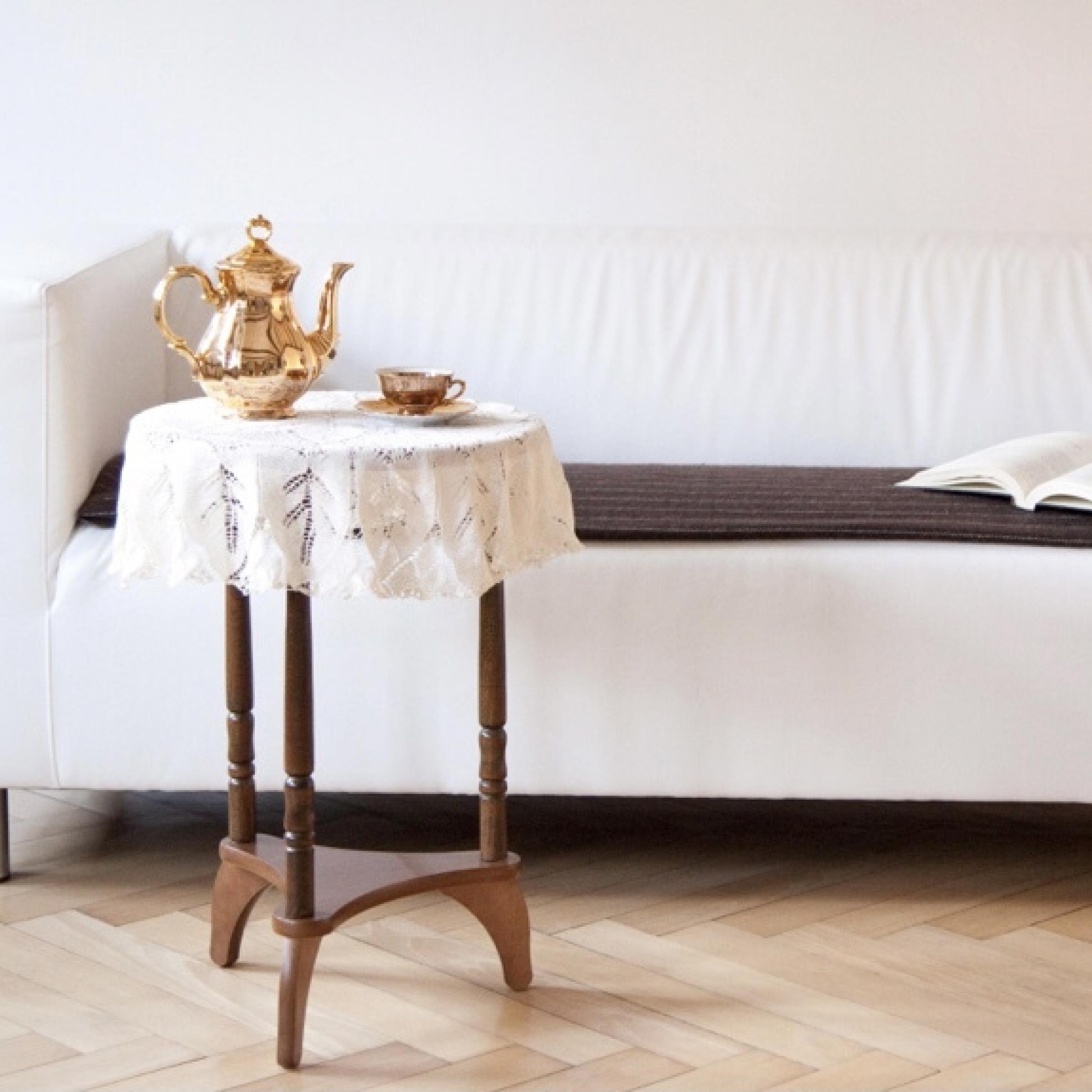 Das Foto zeigt ein weißes Sofa, auf dem ein aufgeschlagenes Buch liegt. Vor dem Sofa steht ein rundes Holztischchen, auf dem eine goldene Kaffeekanne und eine goldene Tasse stehen. Das Bild strahlt Ruhe aus und lädt zum Innehalten ein.