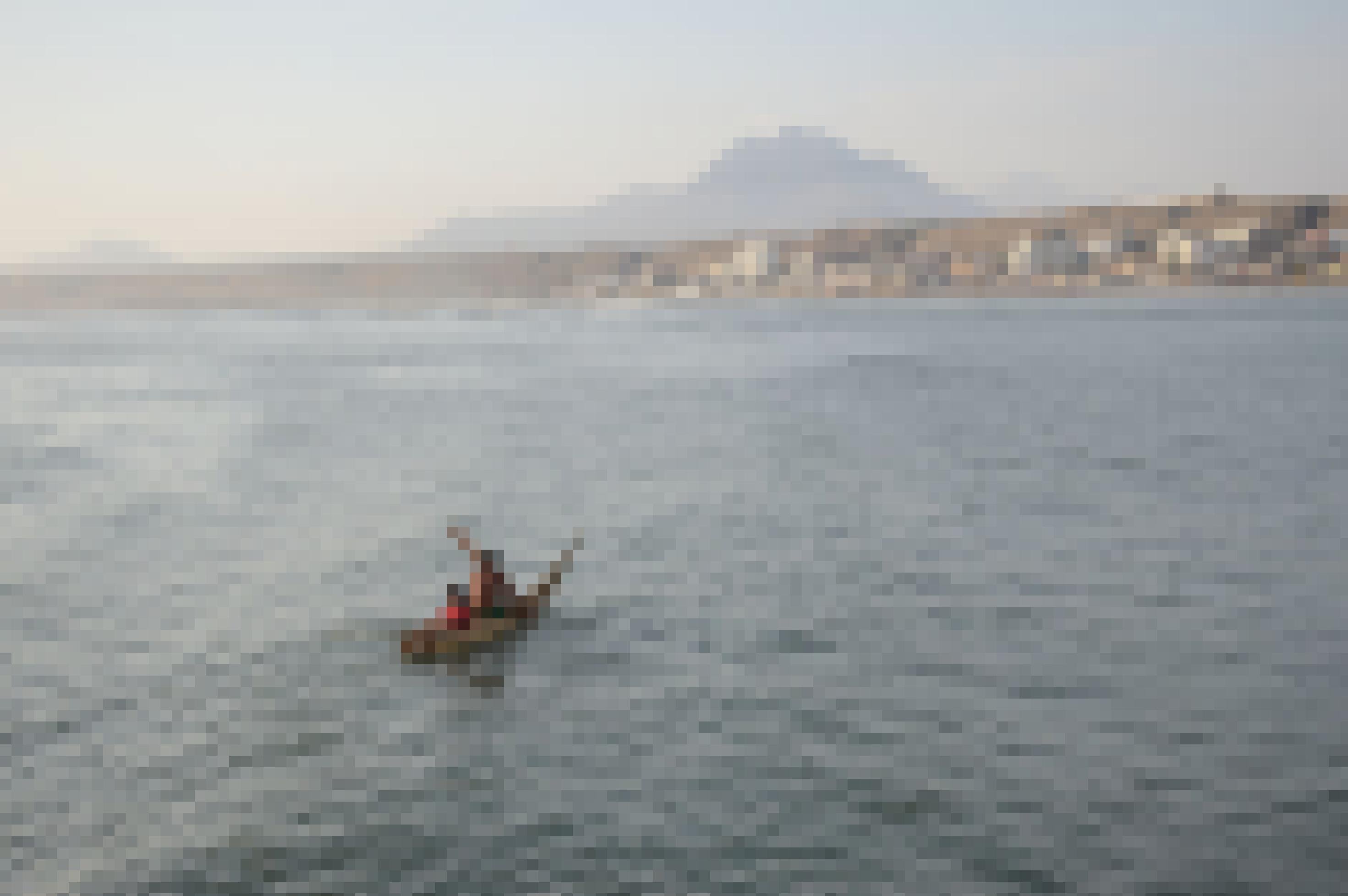 Blick vom Meer auf eine Küste. In der Mitte des Wassers ein Schilfboot, auf dem ein paddelnder Mann und ein Kind sitzt.
